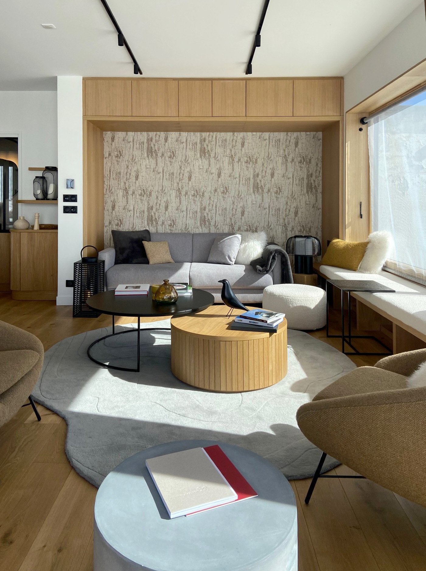 Salon moderne avec canapé gris, chaises en beige, table basse ronde en bois, tapis blanc, mur en texture claire, grande fenêtre avec rideau, décor minimaliste