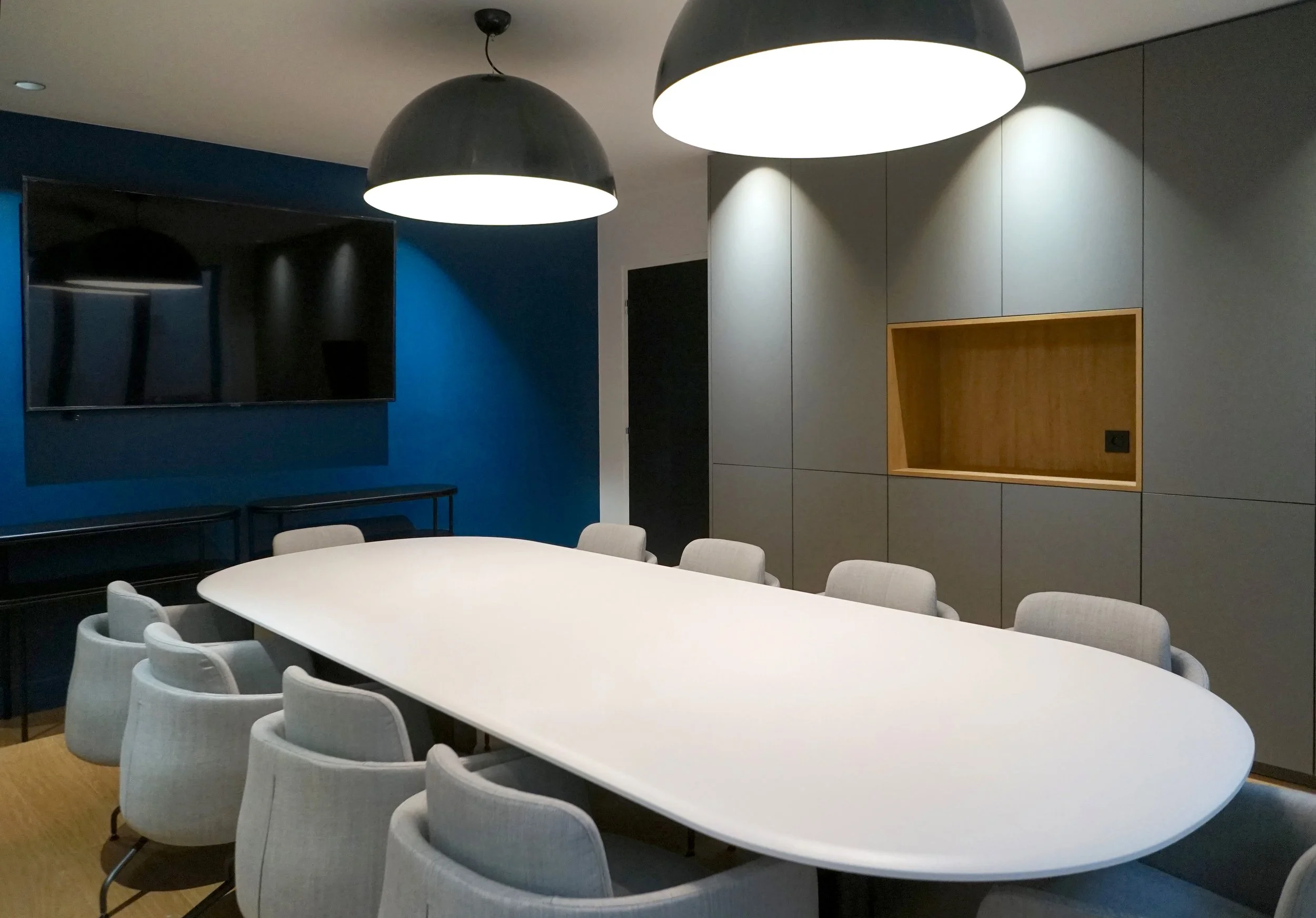 Une salle de réunion moderne avec une grande table ovale blanche entourée de plusieurs chaises en tissu gris clair. Deux grands plafonds ronds noirs avec éclairage blanc diffusent une lumière douce. Un mur bleu foncé à gauche accueille une grande tél