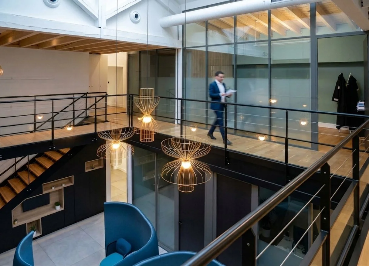Intérieur d'un bureau moderne avec un étage supérieur en verre, des luminaires design en suspension, un homme en costume marchant avec des documents, et des décorations minimalistes.