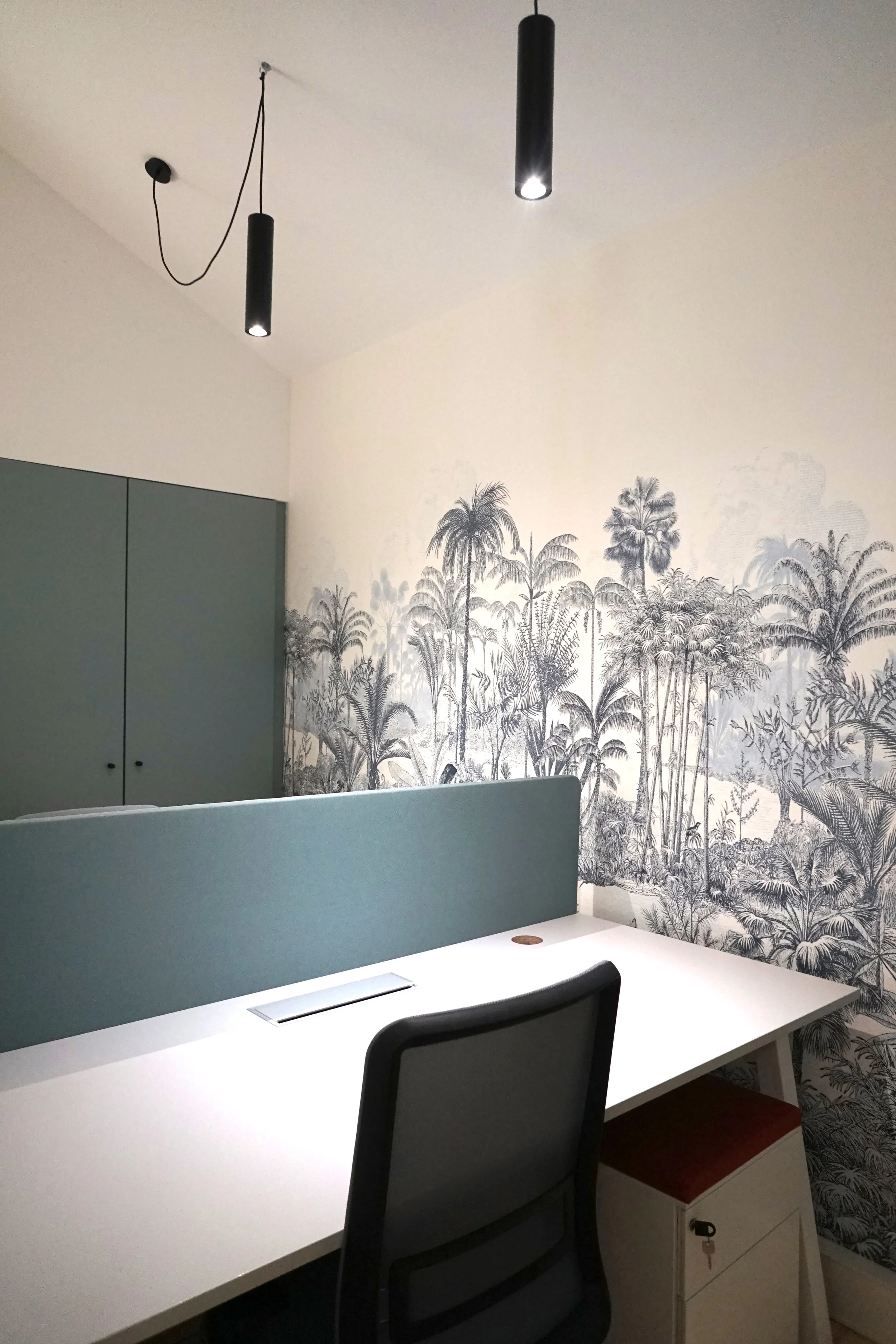 Un bureau moderne avec un mur de coupure en tissu bleu, une chaise ergonomique noir, un bureau blanc avec un panneau de protection en tissu bleu, et un fond mural avec un motif végétal noir et blanc représentant des palmiers et autres plantes tropica