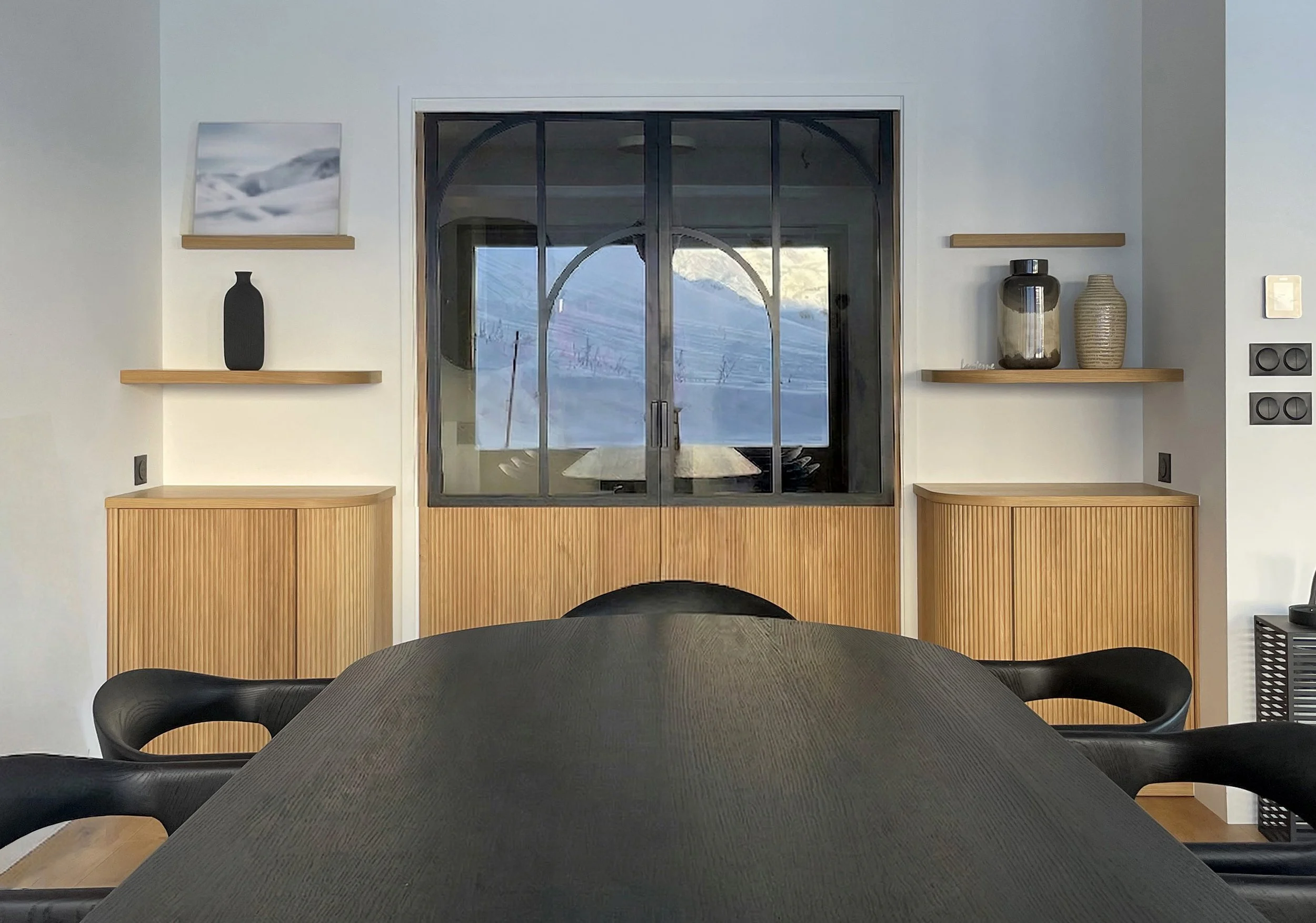 Intérieur moderne avec une table noire, des chaises, une cheminée en bois avec des étagères, et des fenêtres laissant voir un paysage hivernal avec des montagnes et de la neige.