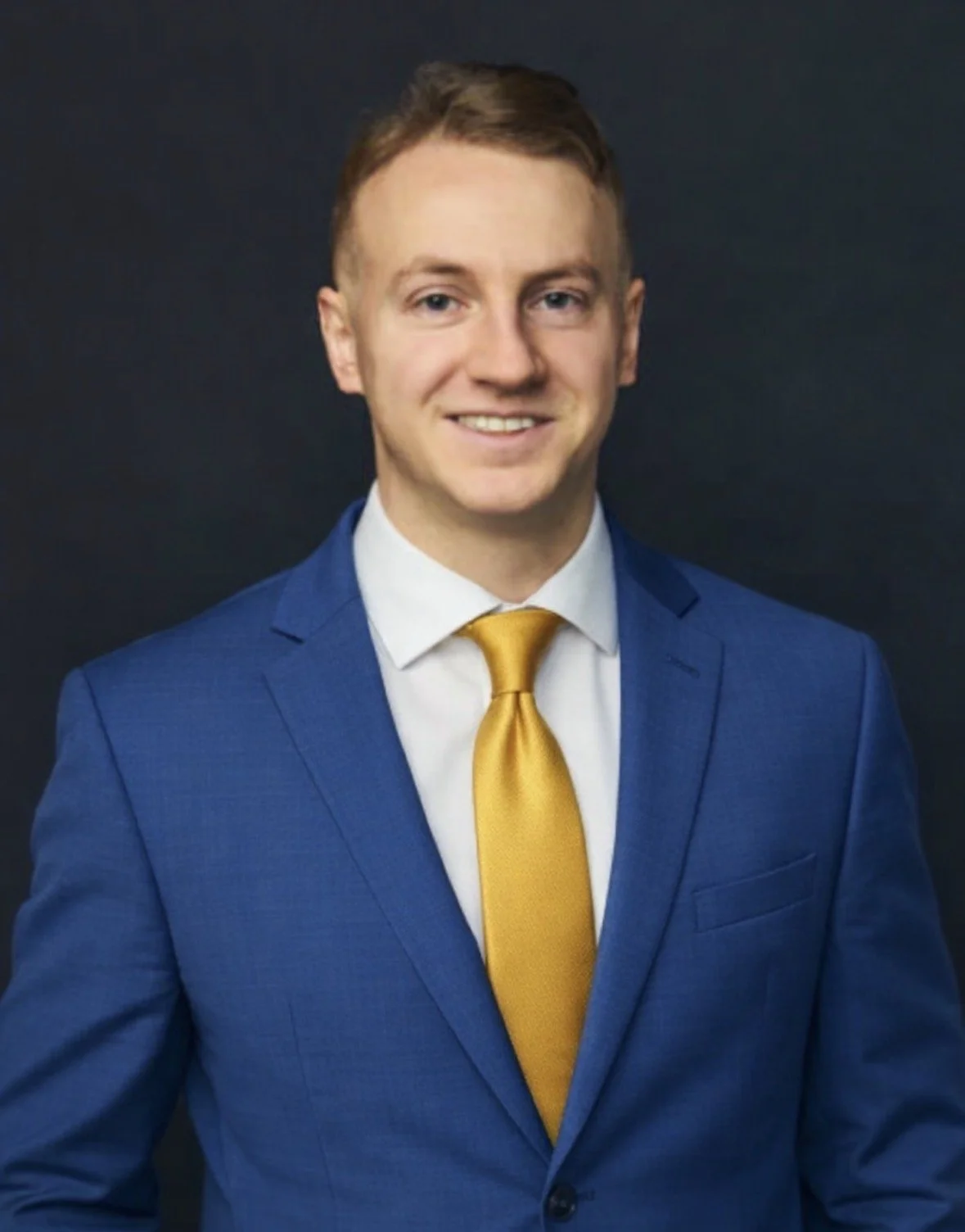 A young man with light skin, short light brown hair, wearing a blue suit, white shirt, and yellow tie, smiling against a dark background.