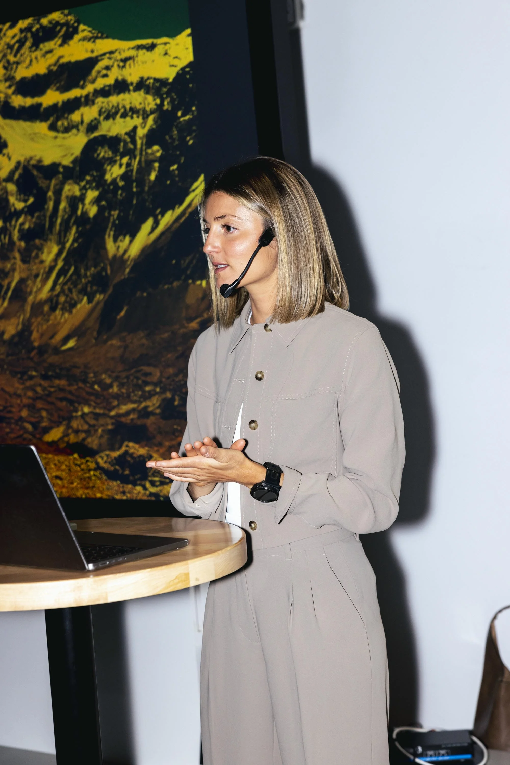 Une femme portant un casque de communication, debout à côté d'un ordinateur portable sur une table en bois, en train de donner une présentation, avec un écran affichant une image de paysage montagneux en arrière-plan.