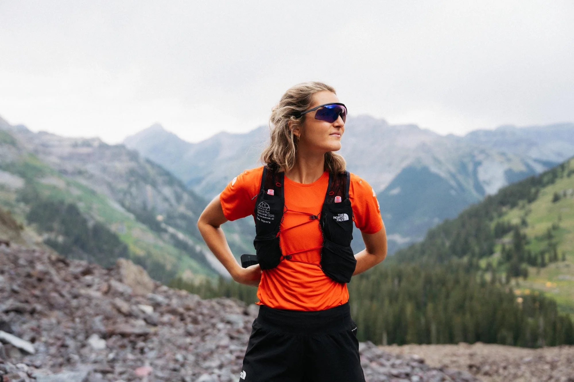 Une femme en vêtements de course à pied orange et noire, portant des lunettes de soleil, se tient dans un paysage de montagne.