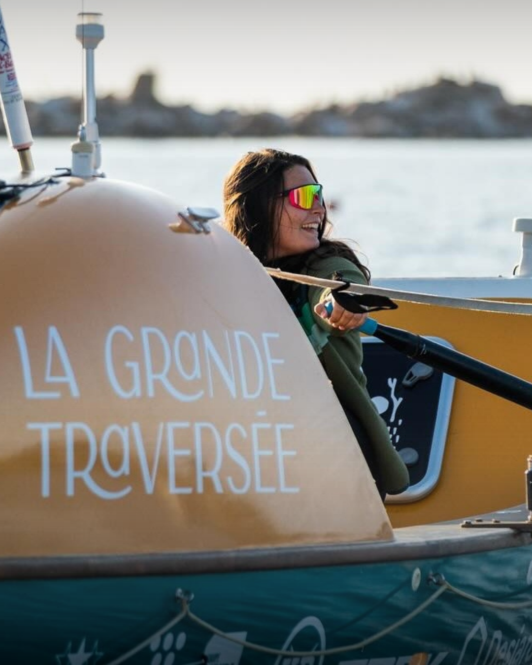 Une femme souriante portant des lunettes de soleil réfléchissantes, assise dans un bateau avec l'inscription 'LA GRANDE TRAVERSÉE'.
