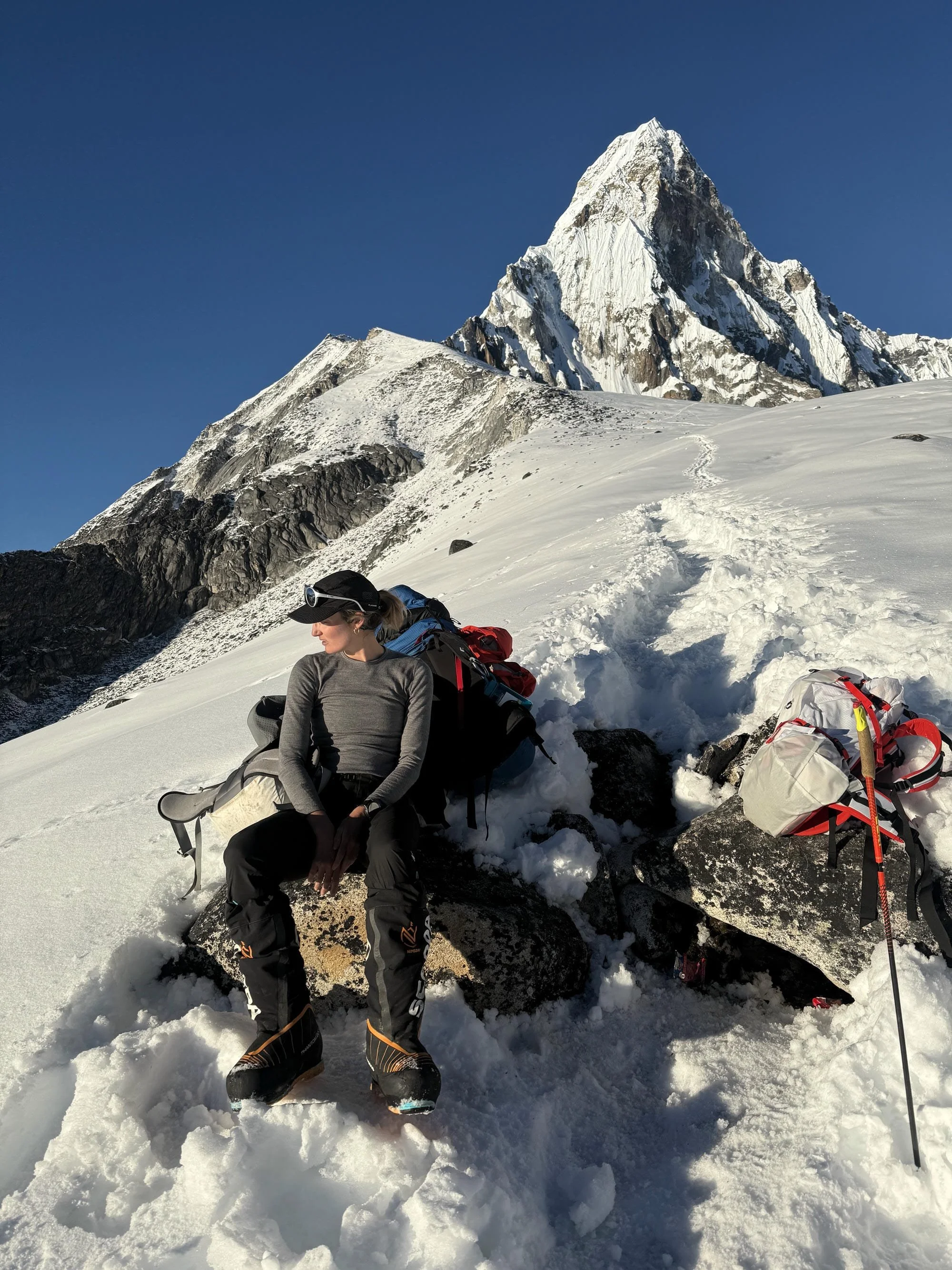 Une femme équipée pour l'alpinisme, assise sur un rocher dans la neige, avec un paysage de montagnes enneigées en arrière-plan.
