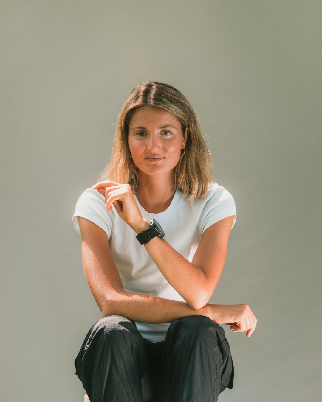 Jeune femme aux cheveux bruns, portant un t-shirt blanc et un pantalon noir, assise contre un fond gris, regardant la caméra, avec une montre noire à son poignet gauche.