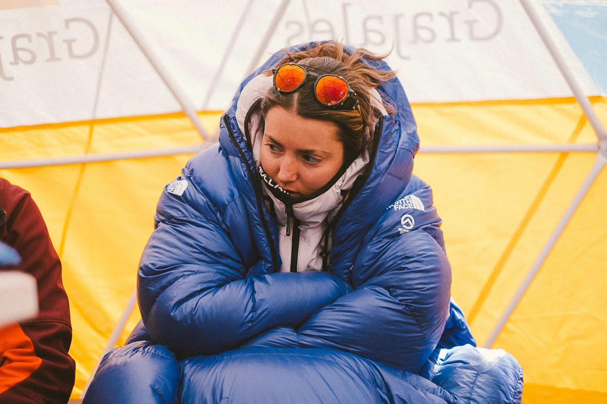 Une femme portant une veste bleue avec des lunettes de soleil à miroir orange, assise dans un endroit probablement en extérieur avec une tente jaune en arrière-plan.
