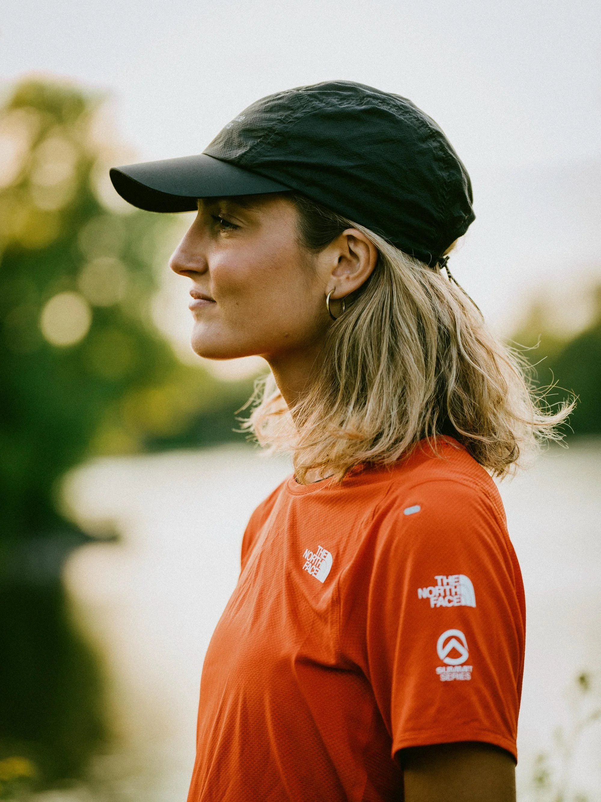 Profil d'une femme portant un chapeau noir et un t-shirt orange de la marque The North Face, avec plusieurs logos sur la manche.