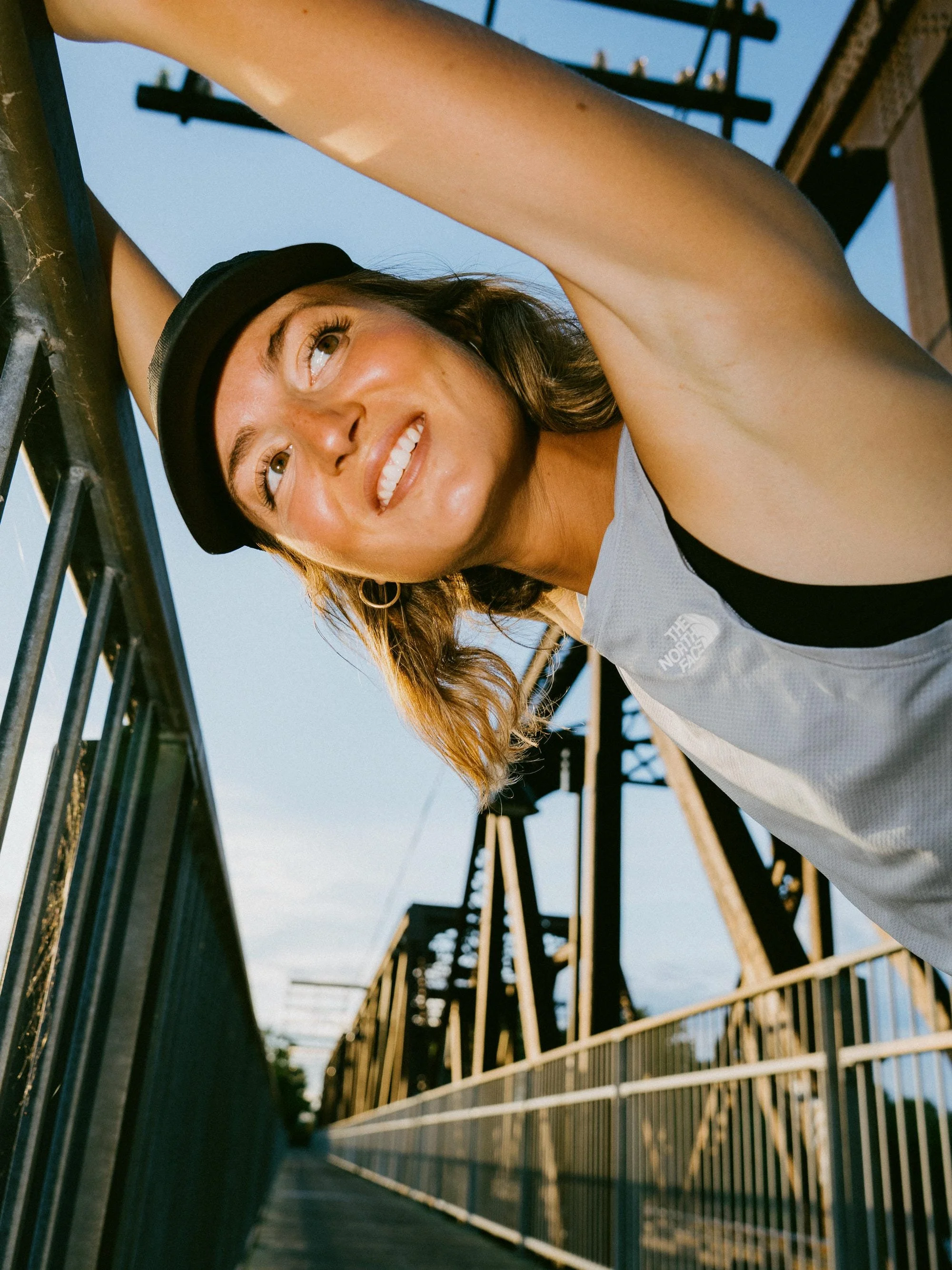 Une jeune femme portant une casquette noire regarde vers le haut en souriant, dans un pont en métal au coucher du soleil.
