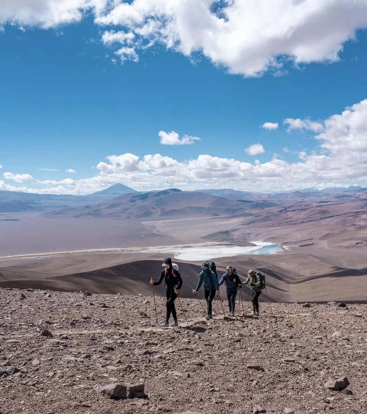 Groupe de quatre randonneurs avec des sacs à dos marchant sur un terrain rocheux dans un paysage désertique avec des montagnes en arrière-plan et un ciel bleu avec des nuages blancs.