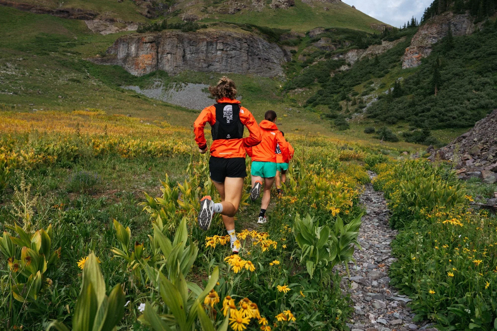 Trois randonneurs portant des vestes orange et des shorts courent à travers un champ de fleurs jaunes dans une vallée entourée de collines verdoyantes et de rochers.