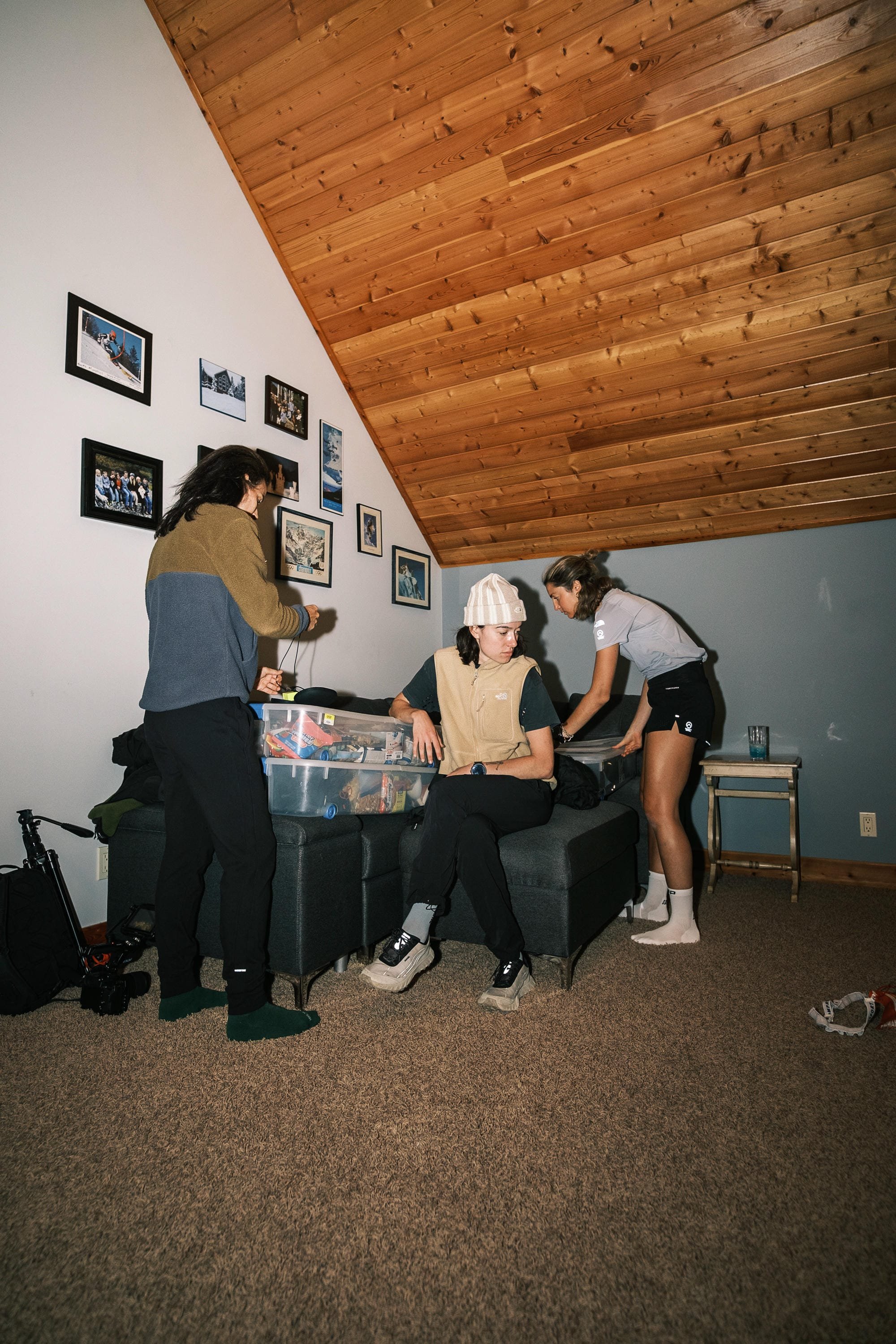 Quatre personnes dans une pièce avec plafond en bois, décorée de plusieurs photos. Une personne en fauteuil regarde vers le haut, deux autres debout discutent près d'une table, une quatrième assise renversée vers eux.