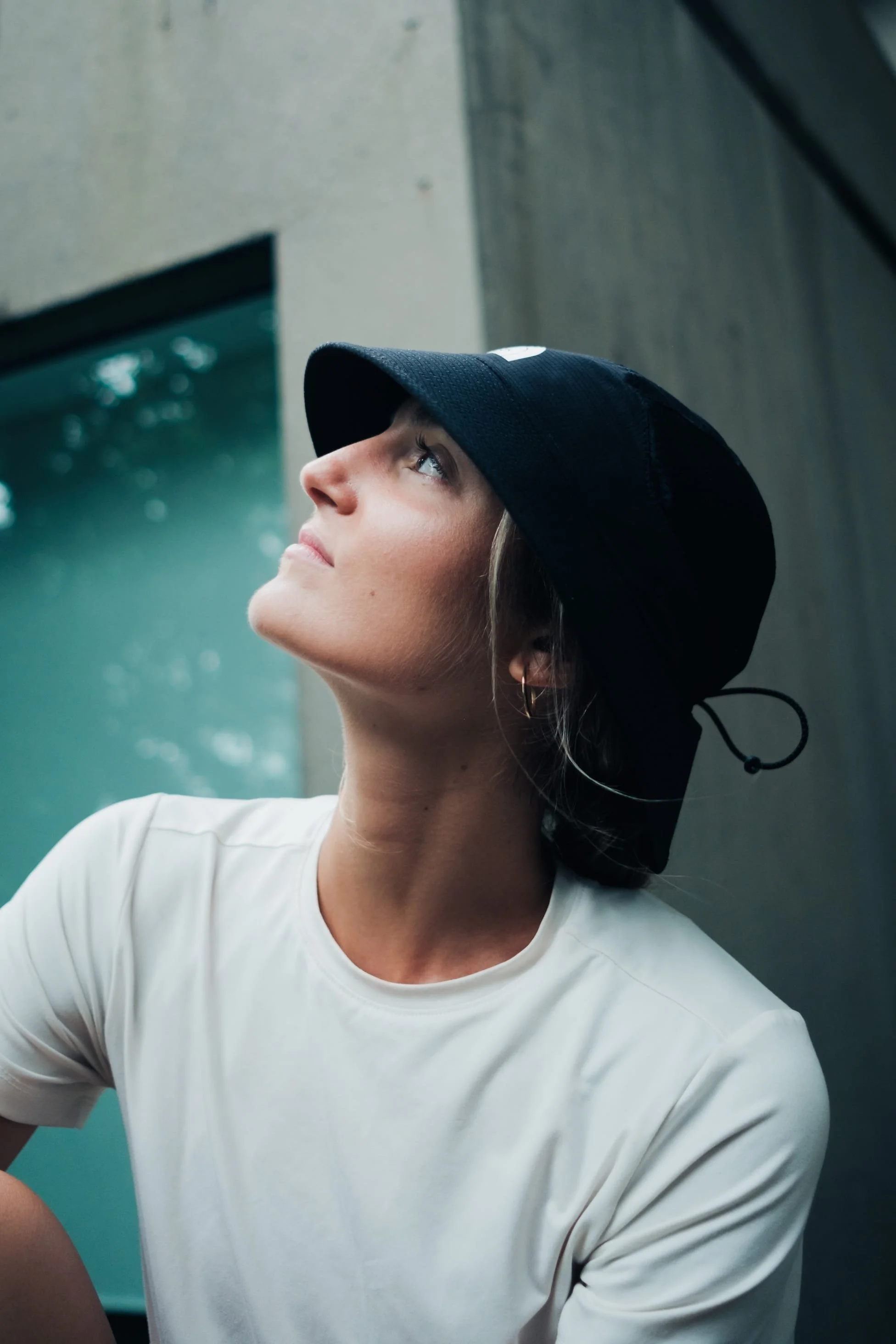 Une femme portant un chapeau noir et un t-shirt blanc regarde vers le haut dans un environnement urbain.