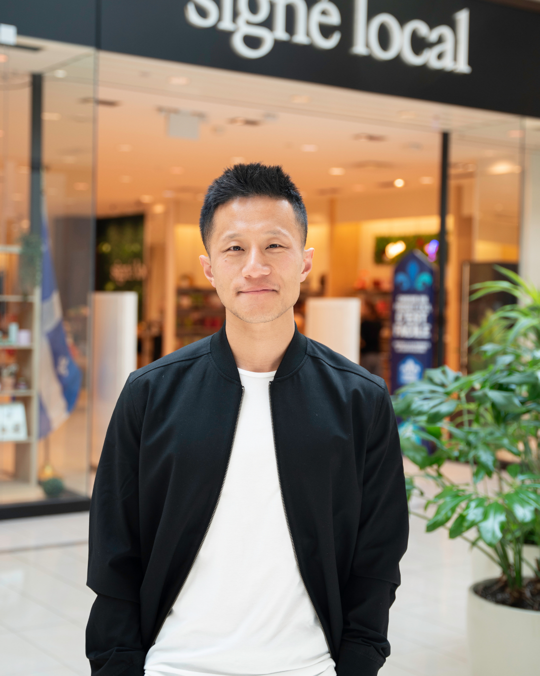 Un homme asiatique souriant portant un bomber noir et un t-shirt blanc, debout devant un magasin avec une enseigne "Signe local", à l'intérieur d'un centre commercial.