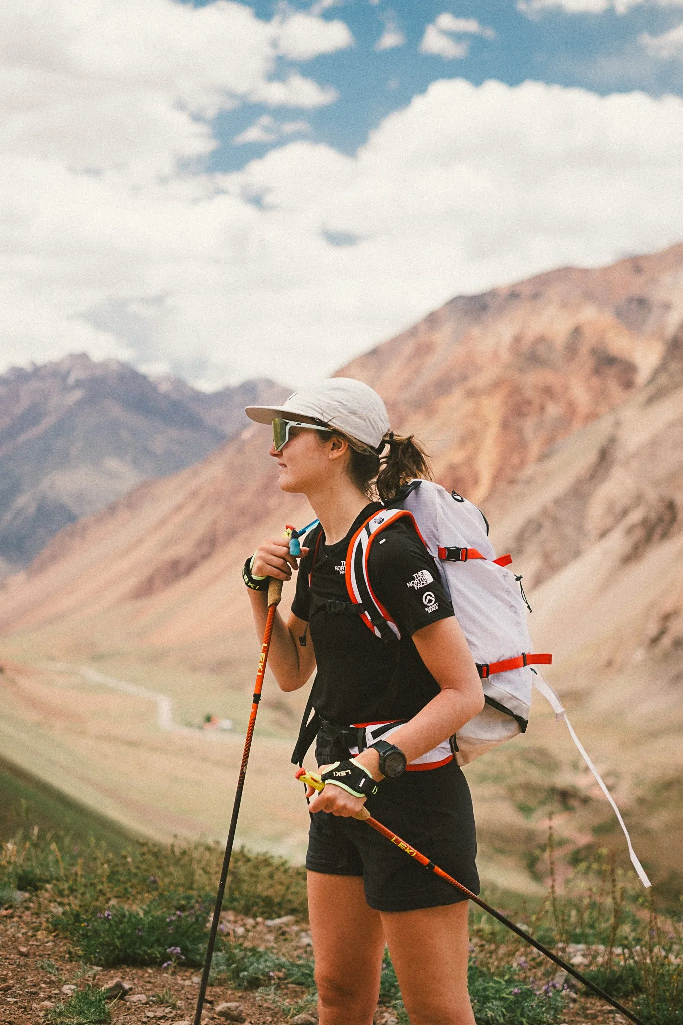 Emmanuel en randonnée dans un paysage montagneux, portant un chapeau, des lunettes de soleil, un sac à dos, une montre, des vêtements de sport, des bâtons de marche, en milieu naturel avec des montagnes et un ciel nuageux.