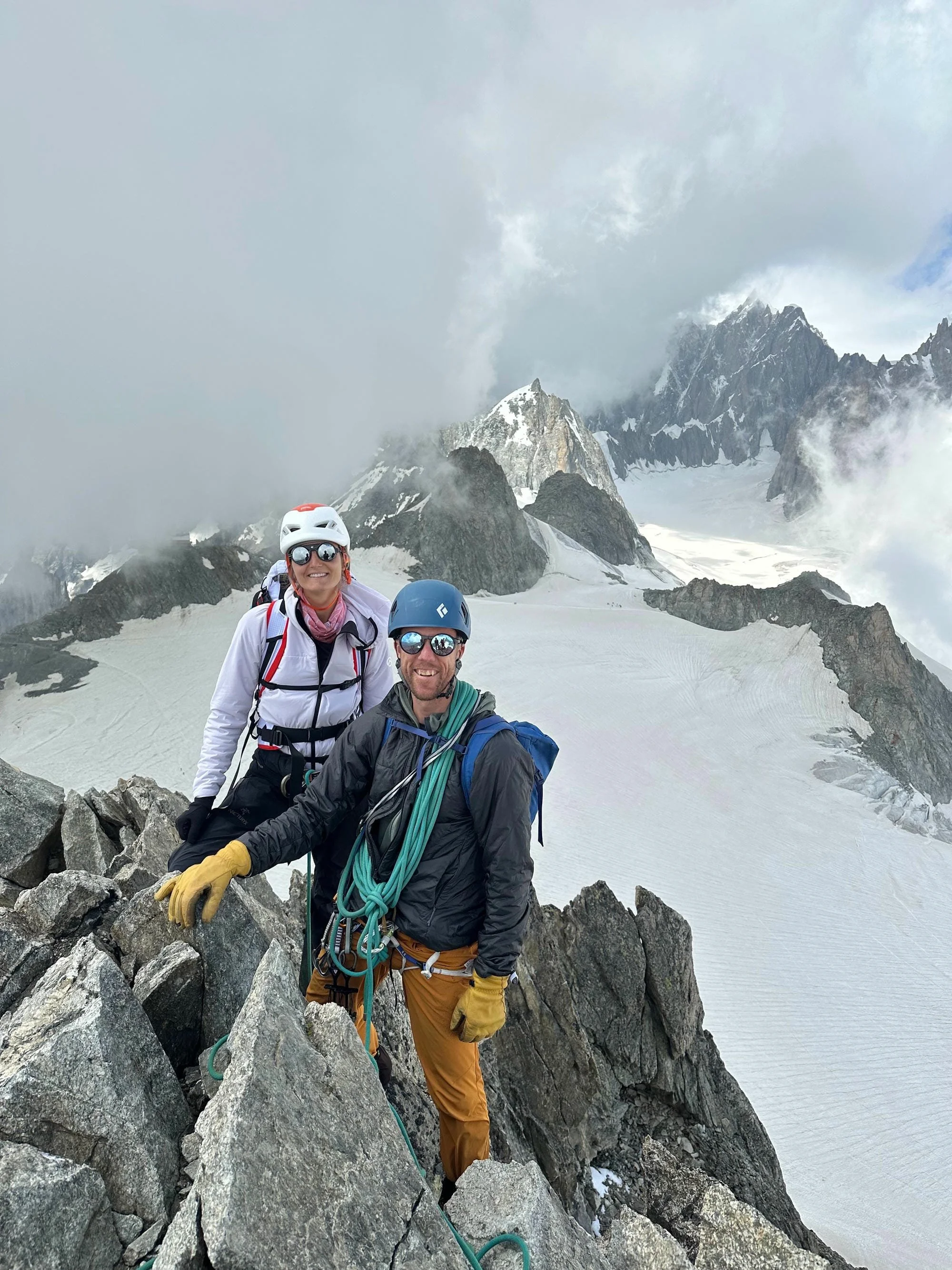 Deux alpinistes en plein effort sur une crête rocheuse enneigée, portant des casques et des équipements d'escalade, avec des montagnes escarpées et des glaciers en arrière-plan