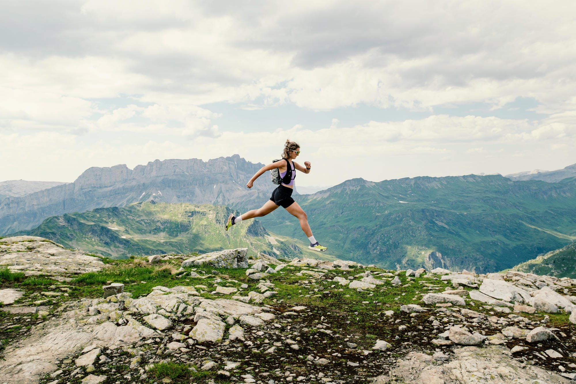 Une femme faisant de la course en montagne sur un sentier rocheux, avec des montagnes en arrière-plan et un ciel nuageux.
