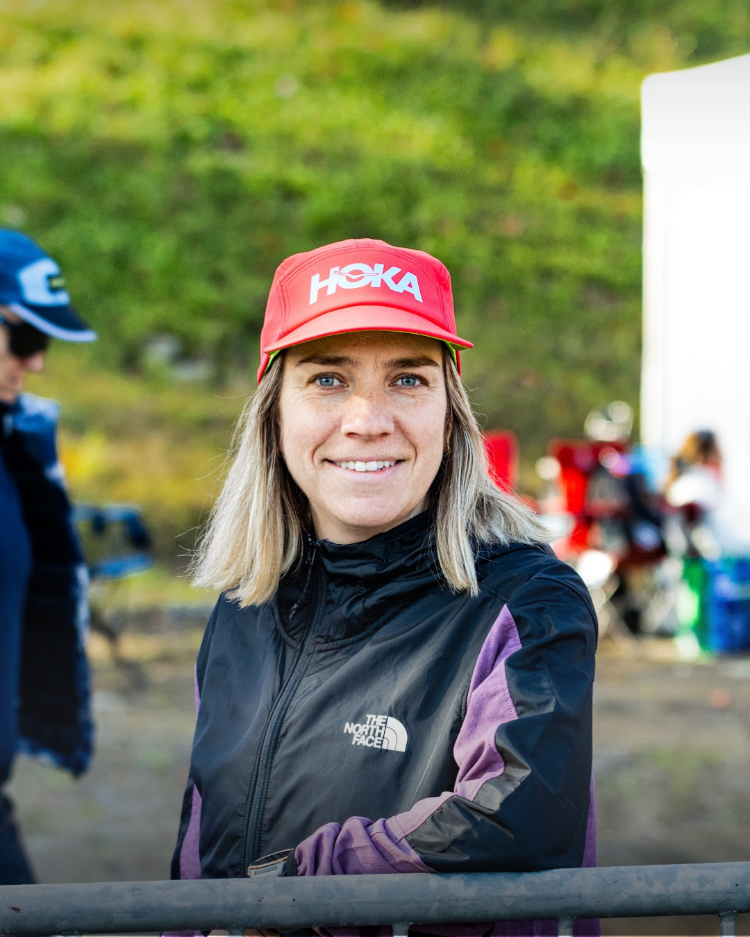 Femme souriante portant une casquette rouge avec le logo HOKA, vêtue d'une veste de sport noire et violette, en extérieur.