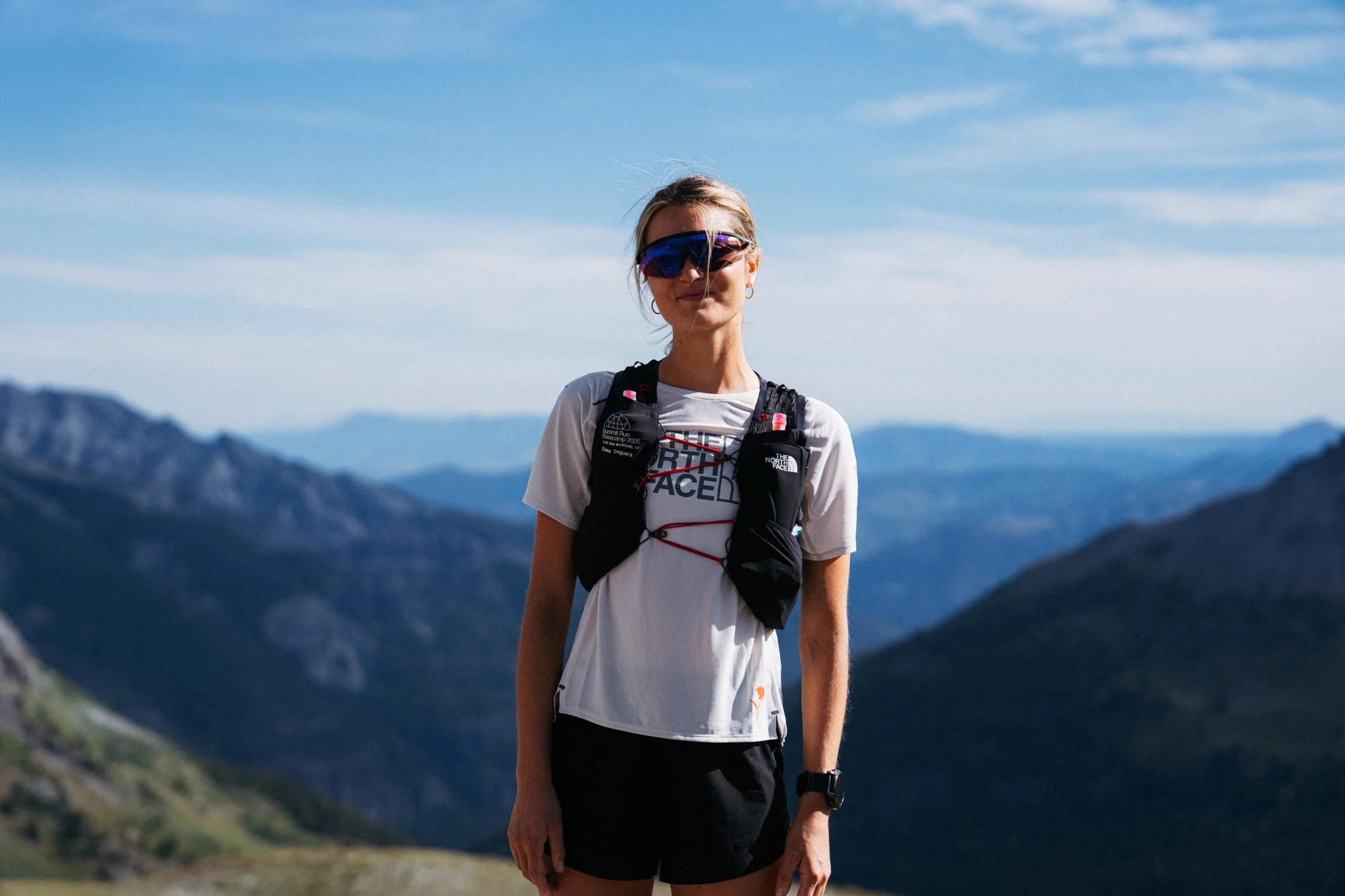 Une femme portant des lunettes de soleil, un t-shirt blanc, un sac à dos et une montre, debout dans un paysage de montagnes avec un ciel bleu et des nuages.