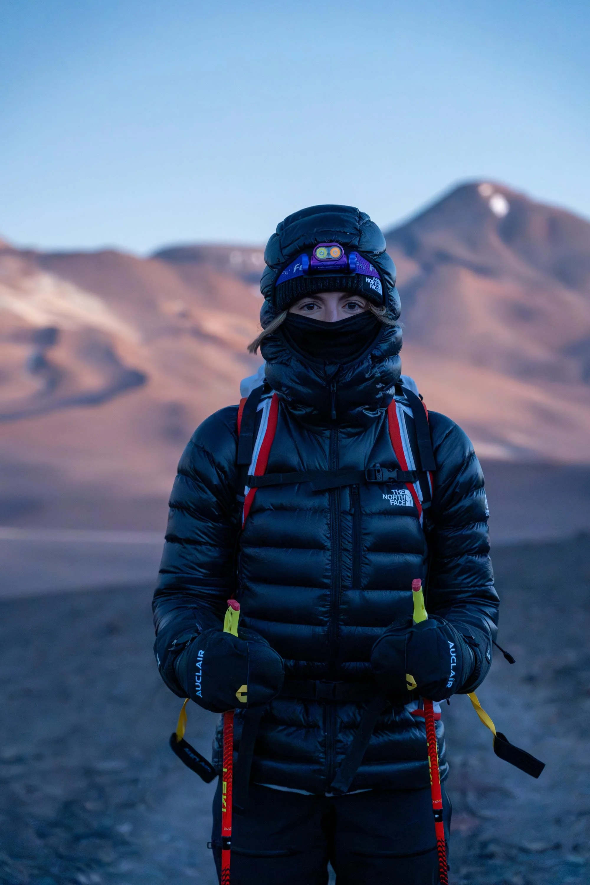 Une personne habillée en équipement de randonnée en montagne, portant une veste noire, un bonnet avec une lampe frontale, un cache-nez noir, et tenant des bâtons de marche rouges et jaunes, avec un paysage de montagnes au coucher du soleil en arrière-plan.