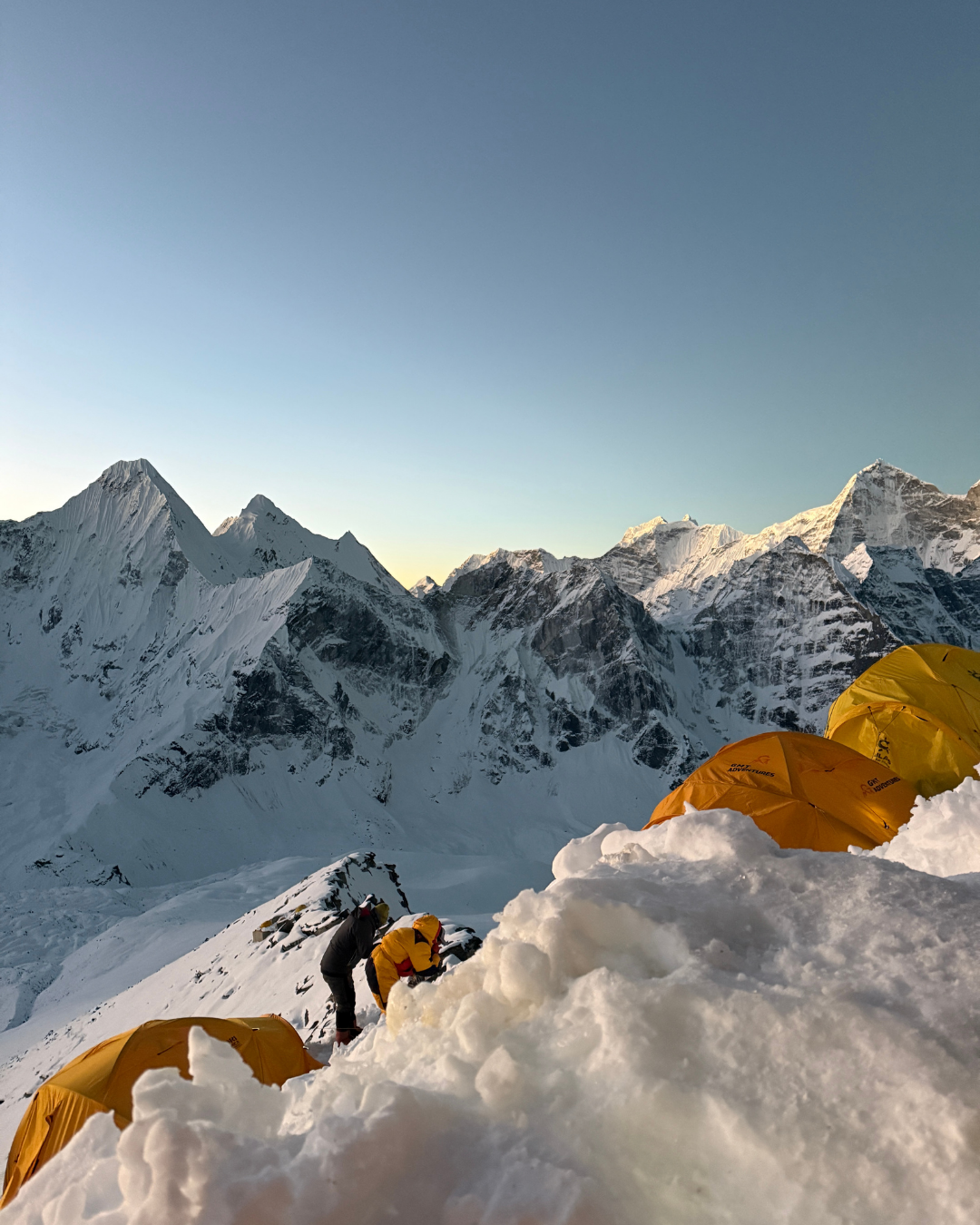 Deux alpinistes en tenue de montagne installent leur campement dans la neige, avec plusieurs tentes orange, dans un paysage de montagnes enneigées au coucher du soleil.