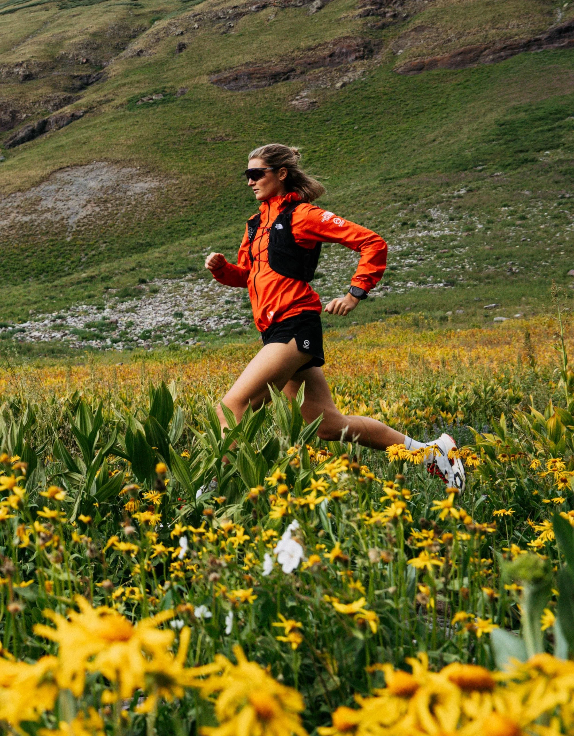 Une femme courent dans un champ de fleurs jaunes, portant une veste orange, des lunettes noires et un sac à dos, avec des montagnes en arrière-plan.