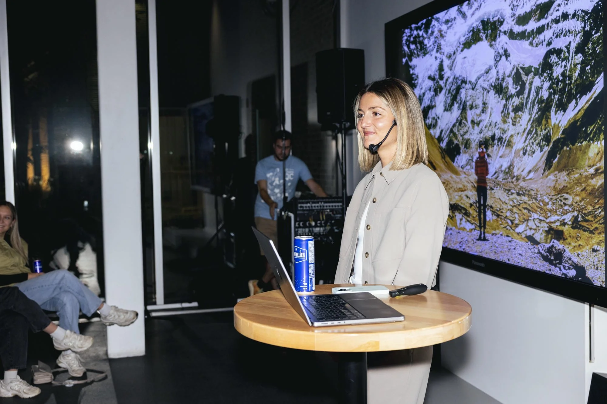Une femme avec un casque microphone se tient devant un grand écran affichant une image de montagnes enneigées, entourée de personnes dans une salle de réunion ou de conférence.