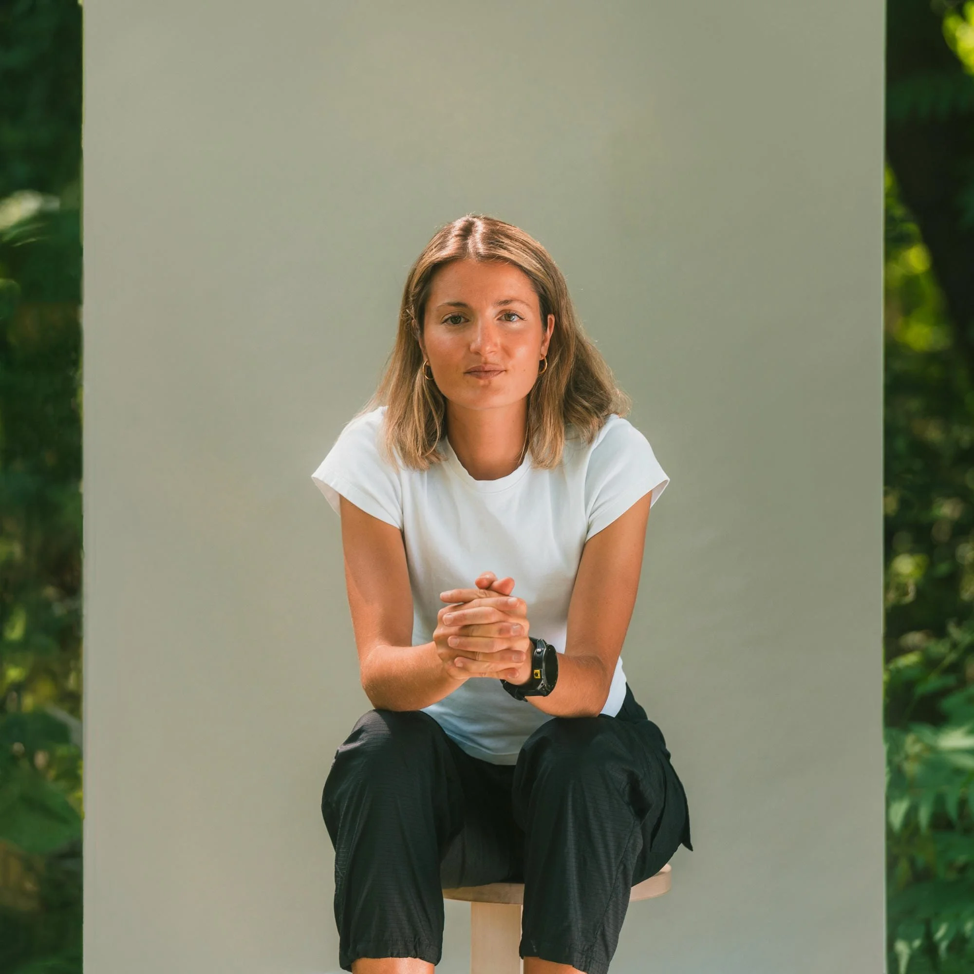 Jeune femme assise sur un tabouret en bois, portant un t-shirt blanc et un legging noir, dans un décor naturel avec des arbres en arrière-plan.