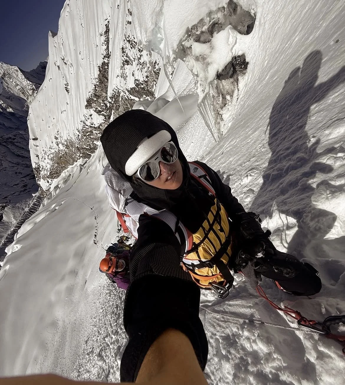 Deux alpinistes équipés de vêtements chauds et de lunettes de soleil escaladent une face enneigée, avec des montagnes enneigées en arrière-plan.