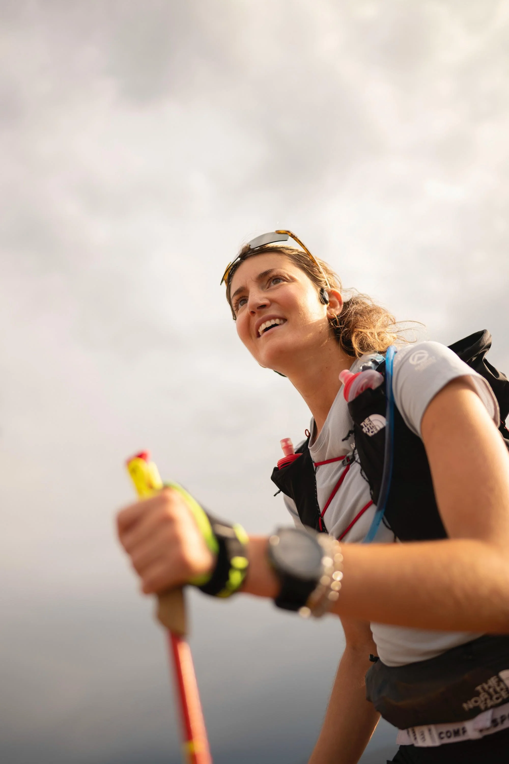 Une femme souriante portant des lunettes de soleil, une veste léger, une montre, et un casque audio, tient un bâton ou un équipement de sport, avec un ciel nuageux en arrière-plan.