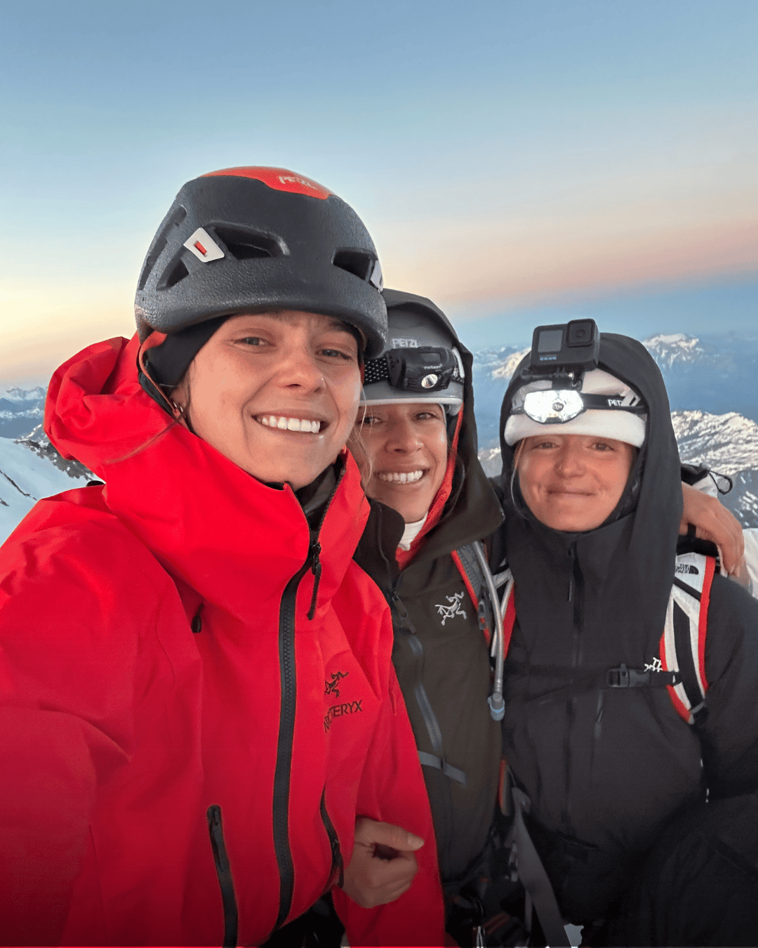 Trois personnes en tenue de montagne avec casques, souriantes, dans un paysage enneigé avec des montagnes en arrière-plan.