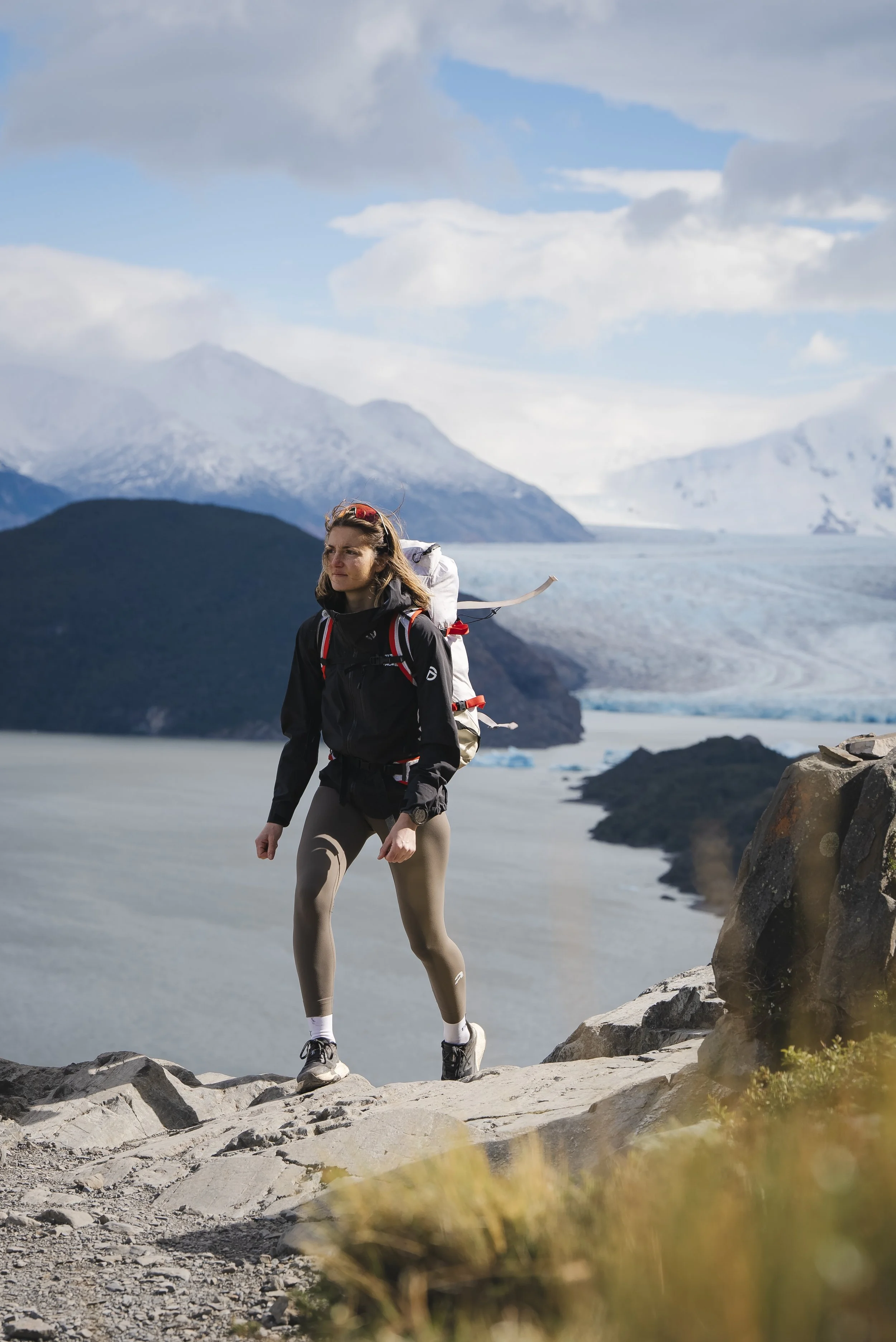 Une femme marche sur un chemin rocheux au bord d'un lac glacé, avec des montagnes enneigées en arrière-plan, sous un ciel partiellement nuageux.