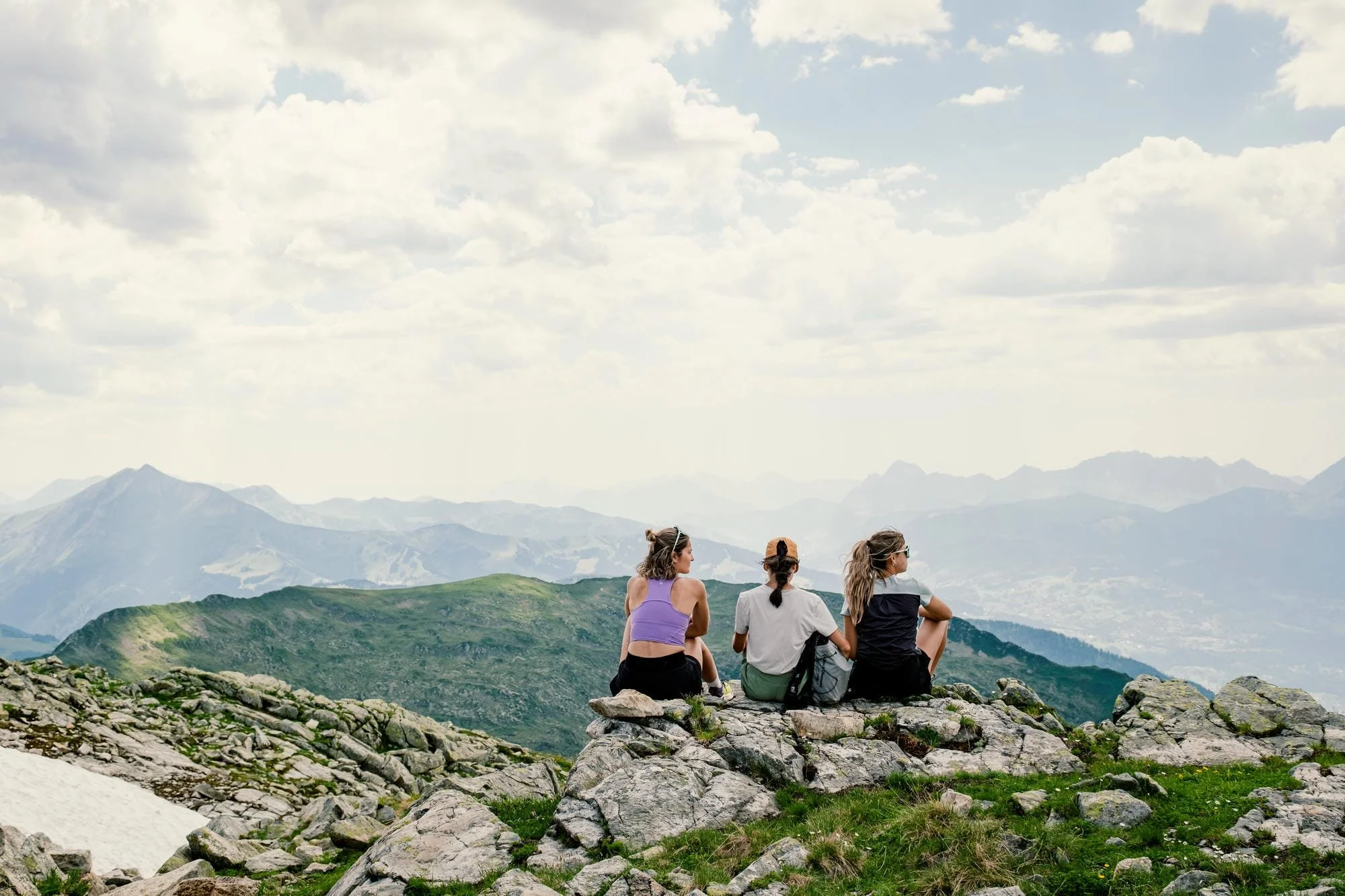 Trois femmes, assises sur une falaise en montagne, regardant un paysage montagneux sous un ciel nuageux.