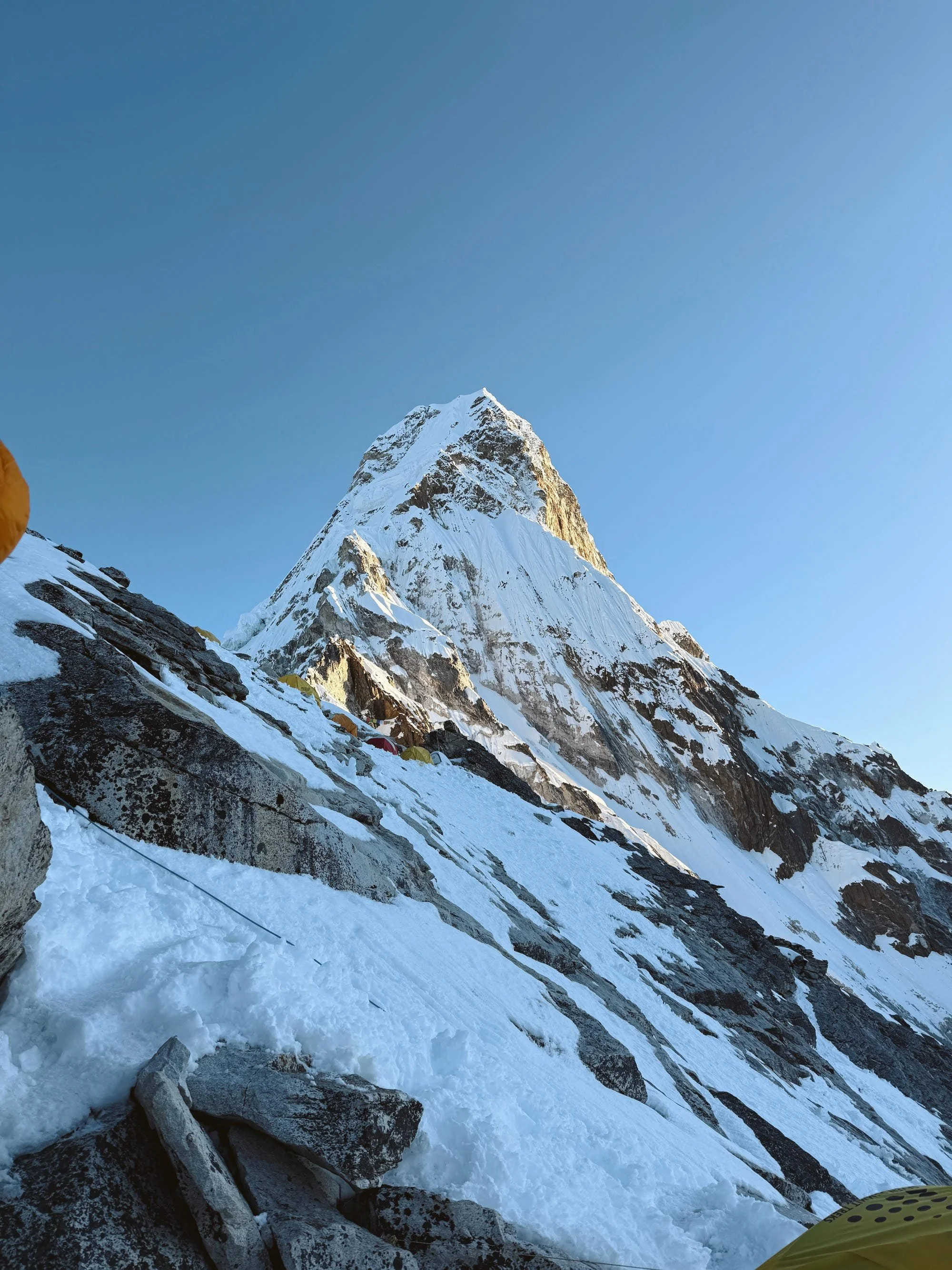 Image d'une montagne enneigée, probablement l'Everest, avec un ciel clair et un terrain glacé en premier plan. À gauche, une personne en équipement d'alpinisme jaune est visible partiellement.
