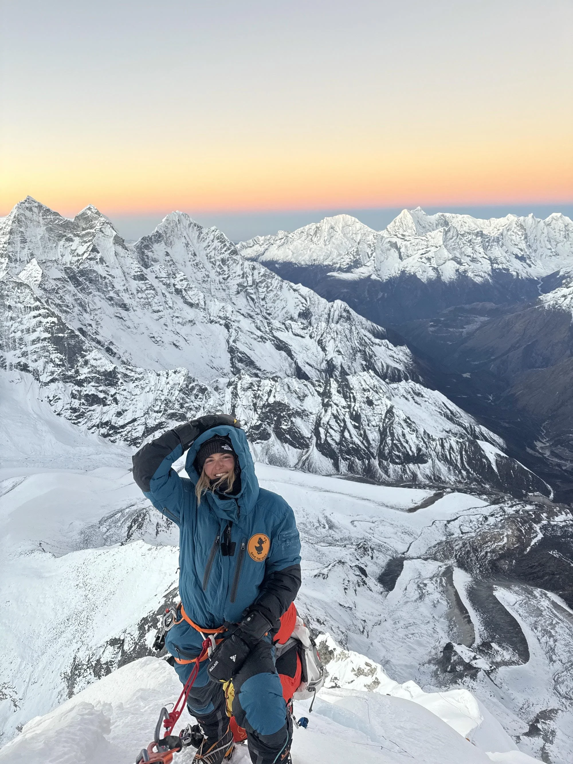 Une femme souriante en tenue de montagne, équipée pour l'alpinisme, pose avec des montagnes enneigées en arrière-plan au lever ou au coucher du soleil.
