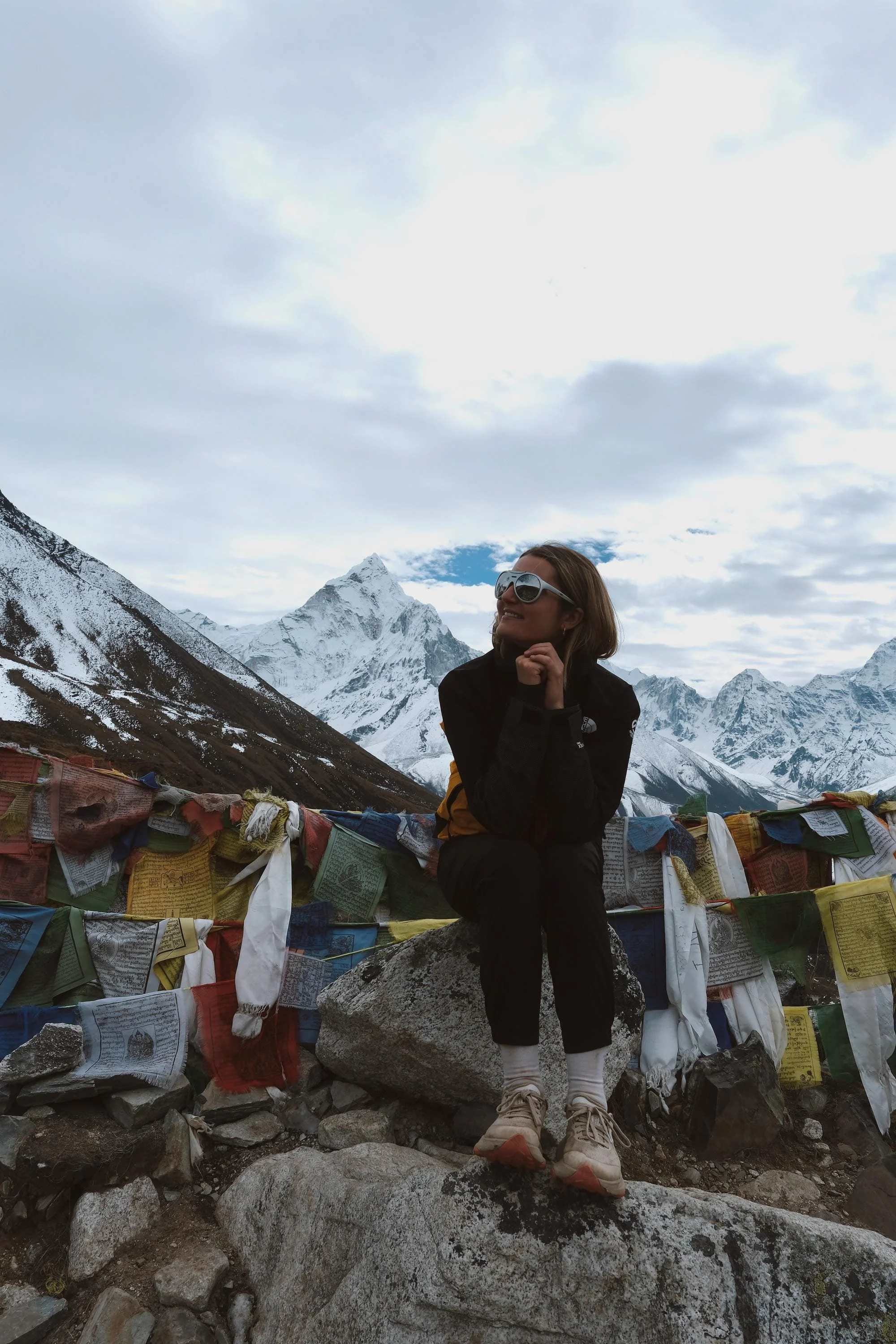 Une femme assise sur une roche dans un paysage de montagnes enneigées, portant des lunettes de soleil, un manteau noir et des chaussures de randonnée, avec un mur de drapeaux de prière tibétains en arrière-plan.