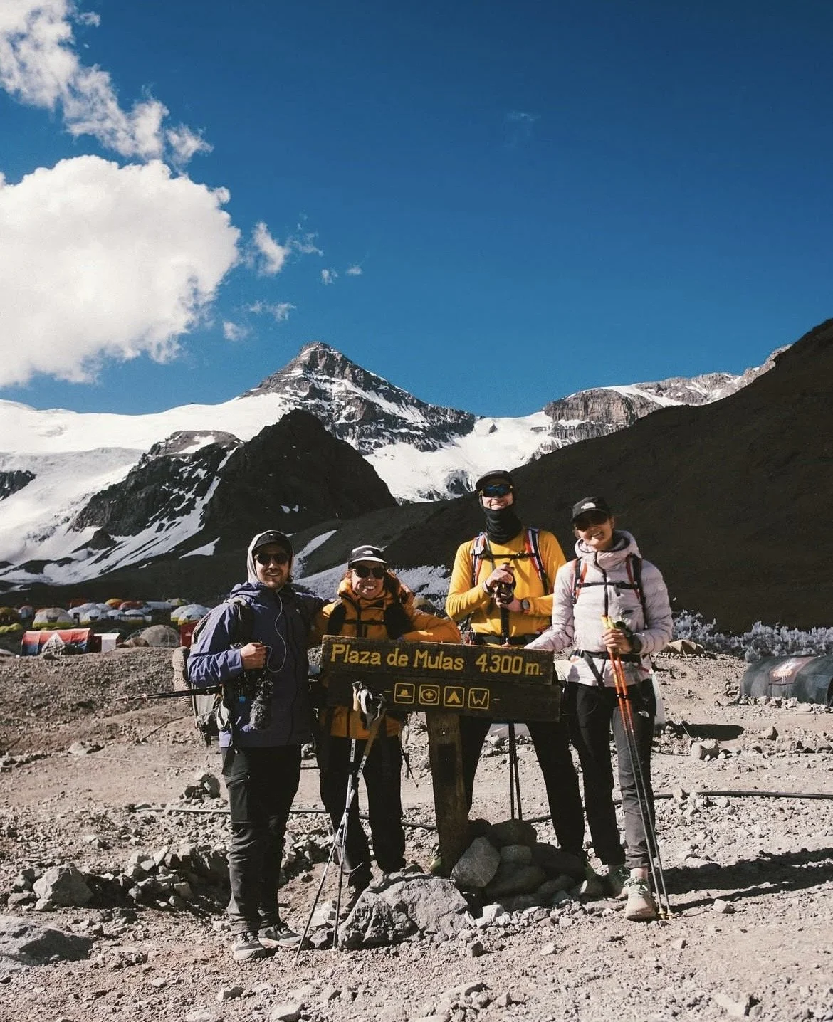 Groupe de quatre randonneurs avec sacs à dos devant un panneau indiquant 'Plaza de Mulas 4300 m', avec des montagnes enneigées en arrière-plan.