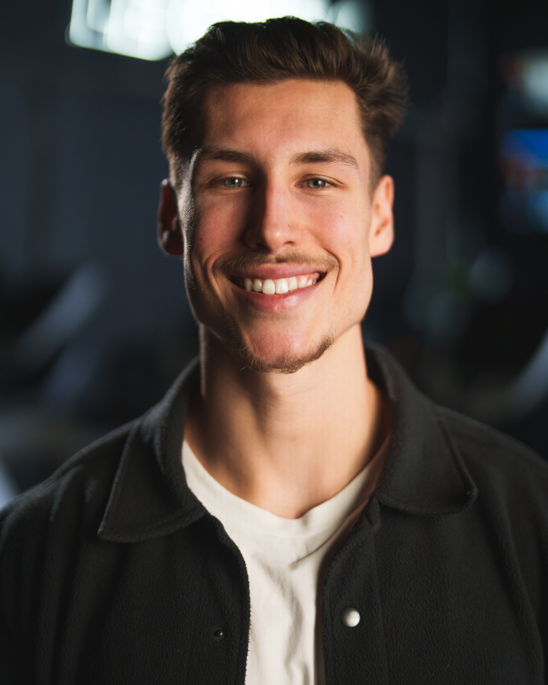 Portrait d'un jeune homme souriant avec une coiffure moderne, portant une veste noire et un t-shirt blanc, dans un environnement intérieur avec un éclairage doux.