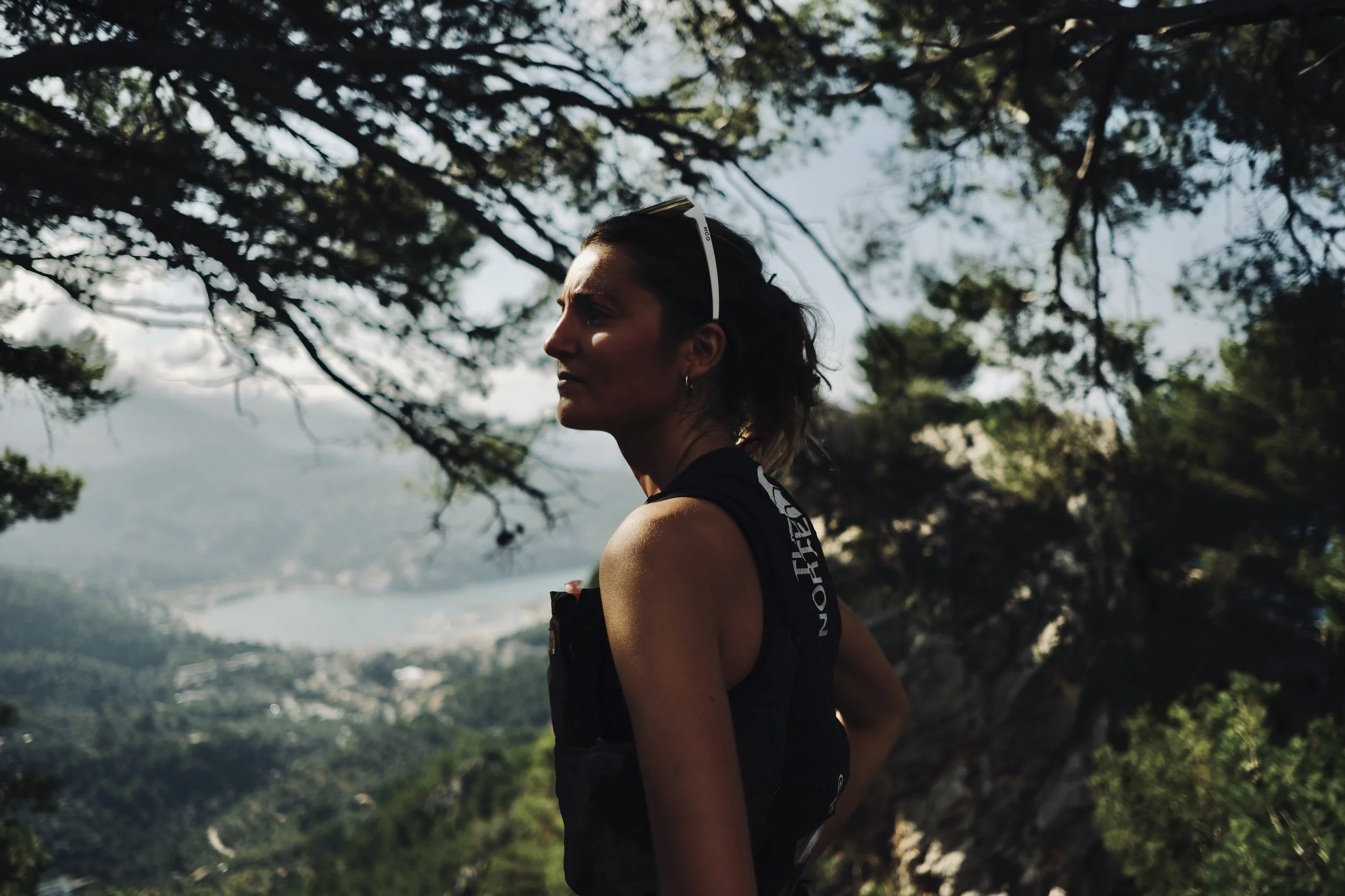 Une femme avec un bandeau blanc portant des écouteurs se tient dans un environnement naturel avec des arbres et une vue sur une vallée en arrière-plan.