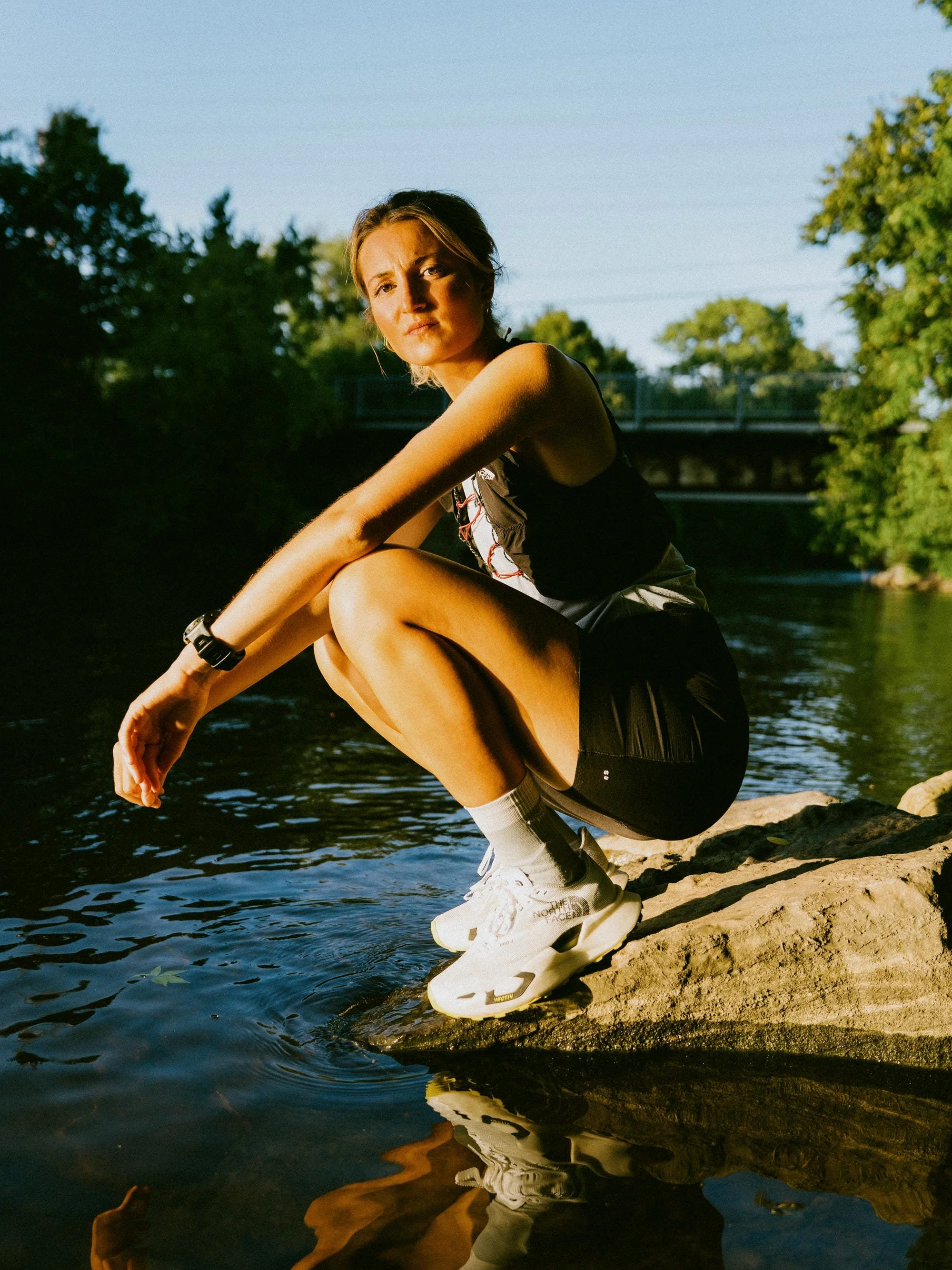 Une femme assise au bord d'une rivière, portant des vêtements de sport blancs et noires, des baskets blanches, et des chaussettes blanches, avec des arbres verdoyants en arrière-plan et un pont en arrière-plan. Elle regarde vers l'objectif avec une expression sérieuse ou pensive.