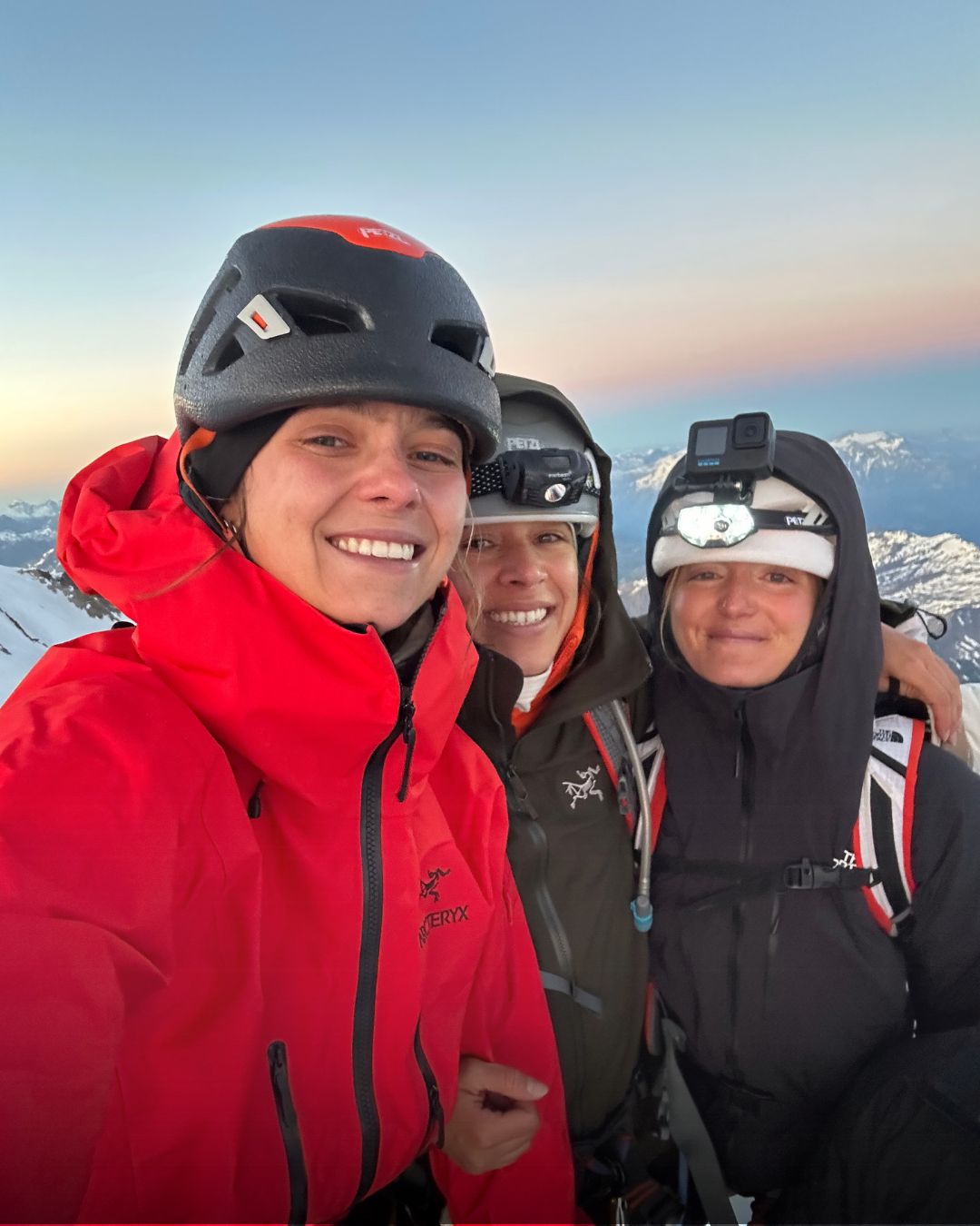 Trois personnes en vêtements de montagne équipées de casques et de lampes frontales, prises avec un sommet enneigé au coucher du soleil.