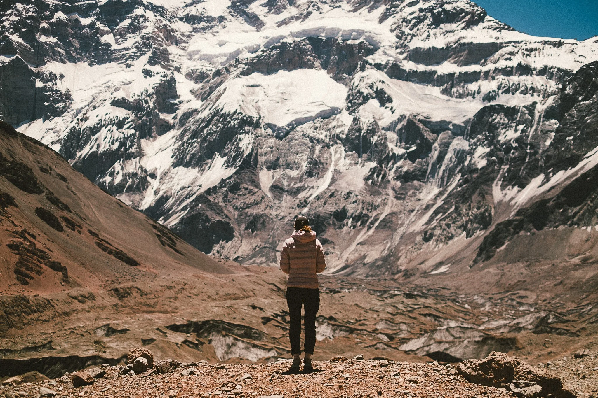 Une personne vue de dos se tient sur un terrain rocheux dans un paysage de montagnes enneigées et de glaciers, avec un ciel bleu clair.