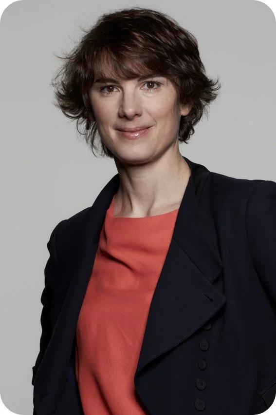 Femme souriante avec cheveux courts, portant une veste noire et un haut orange, contre un fond gris.