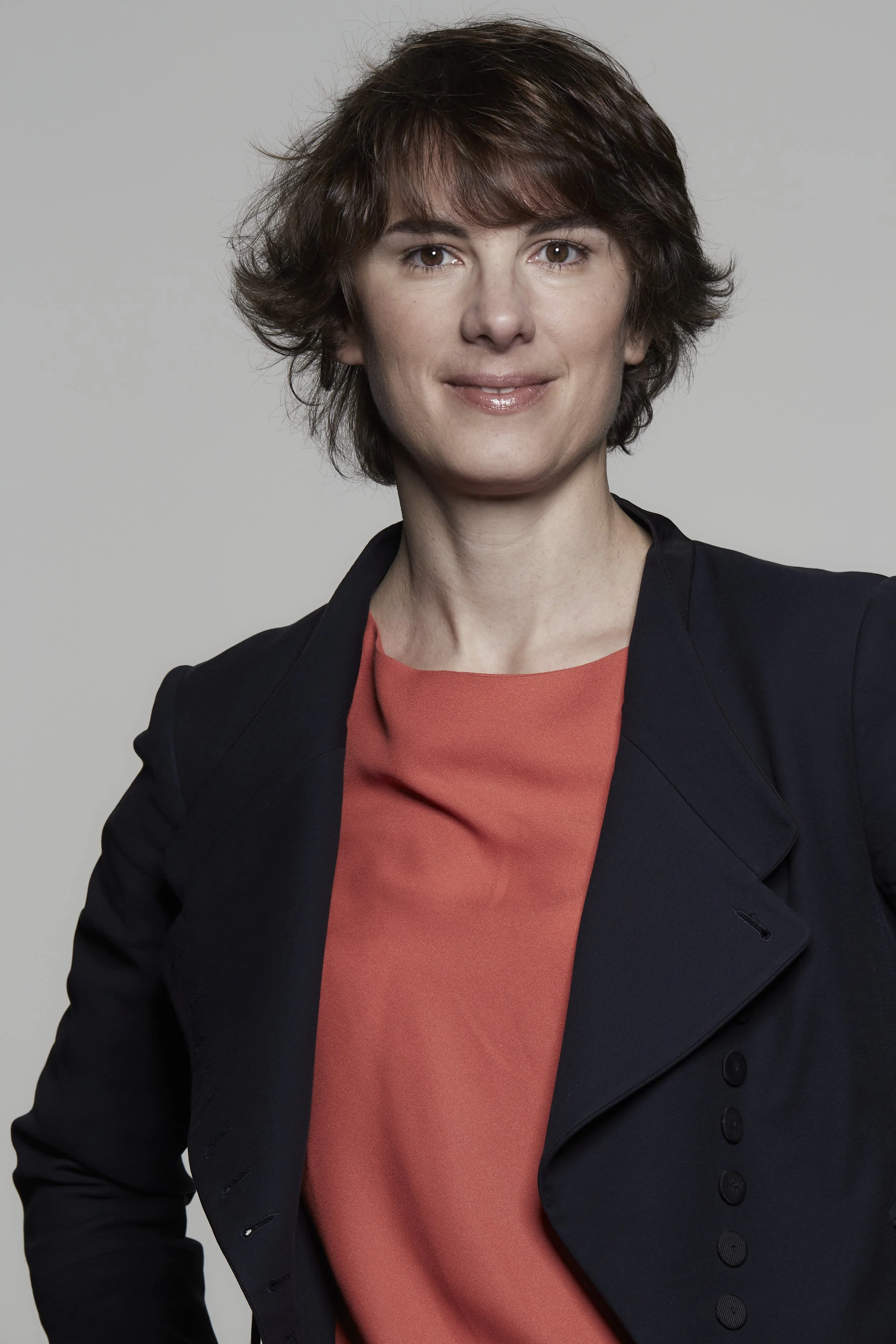 Portrait d'une femme avec des cheveux courts et bruns, portant une veste noire et un haut orange, souriant légèrement, contre un fond gris clair.