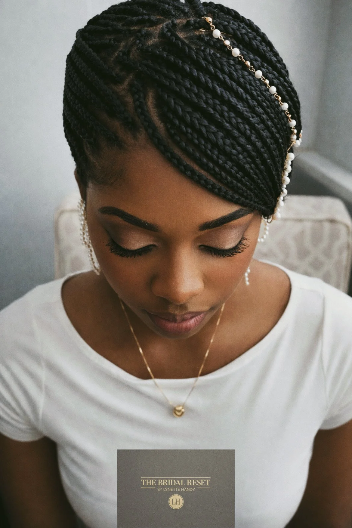 A woman with intricately braided hair adorned with pearl and gold accessories, looking down with her eyes closed, wearing a white top, earrings, and a delicate necklace.