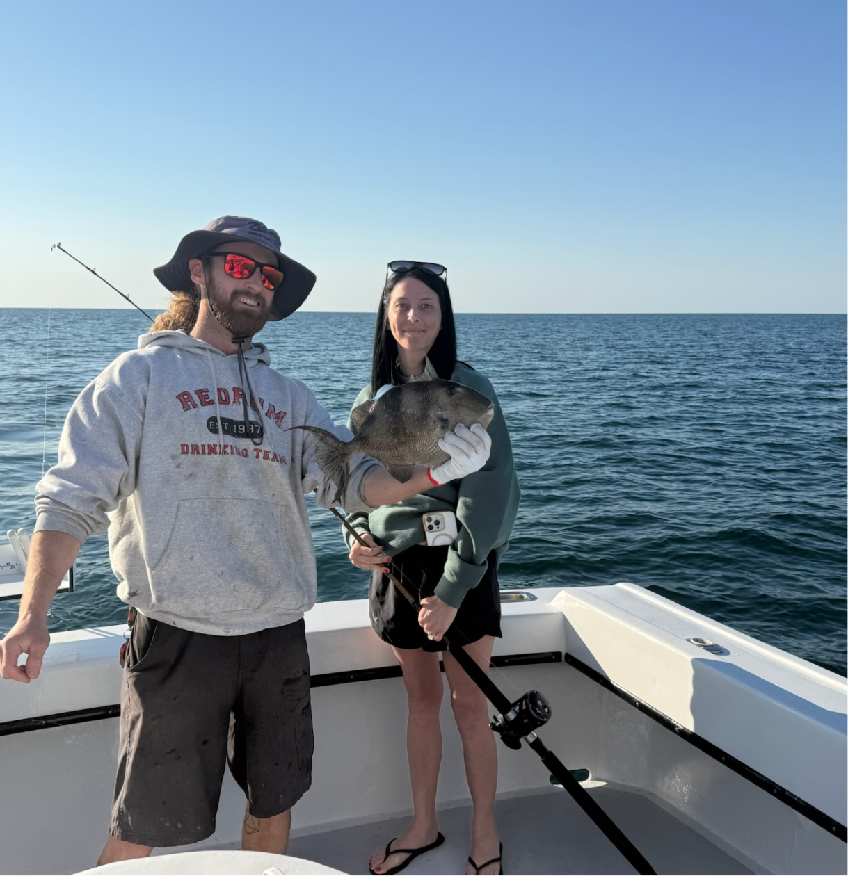 A man and woman stand on a boat in the ocean, holding a fish caught while fishing. The man is wearing a wide-brimmed hat, sunglasses, a hoodie, and shorts, smiling. The woman has long dark hair, sunglasses on her head, a sweatshirt, and shorts, smiling at the camera.