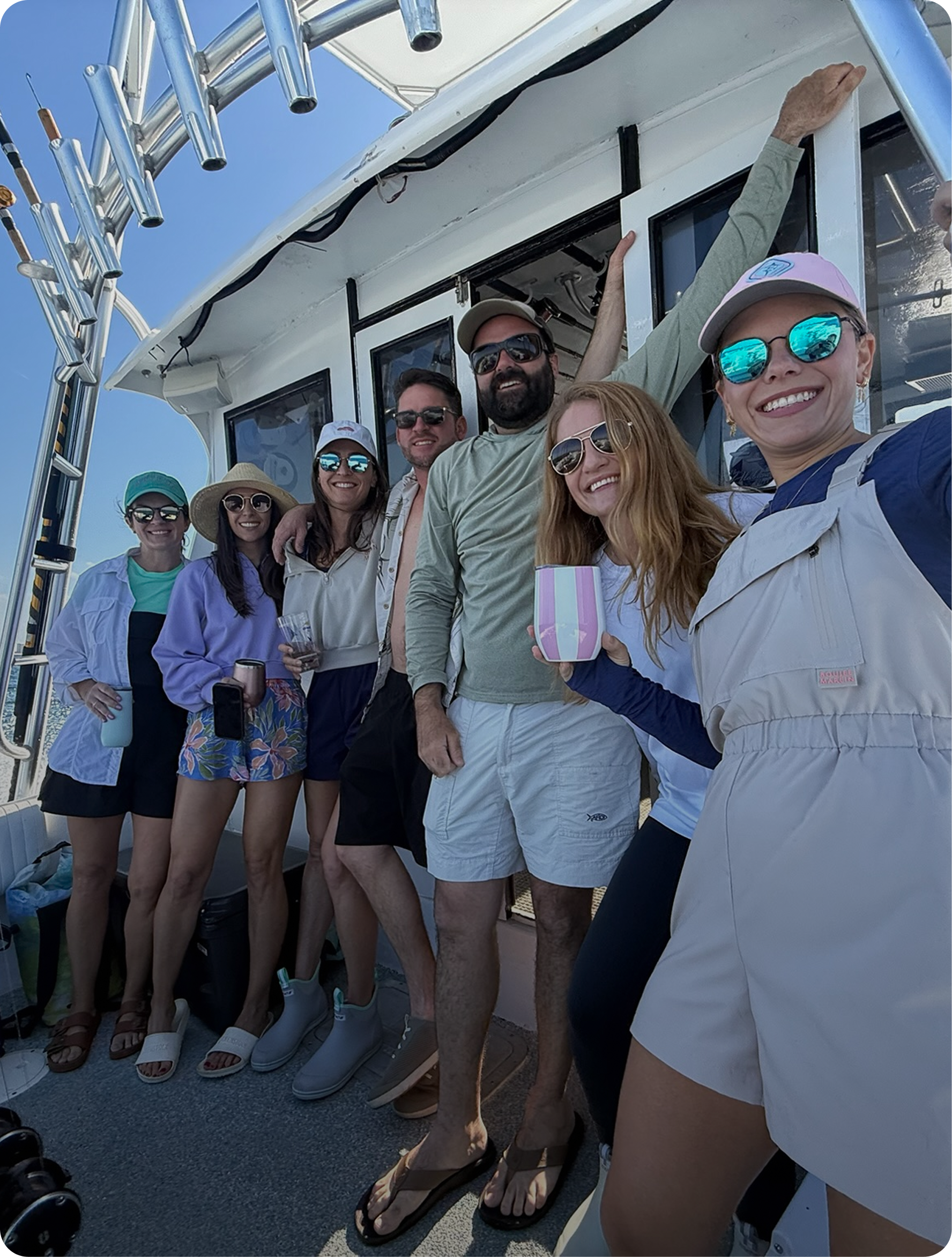 A group of seven friends smiling and posing for a photo on a boat against a bright blue sky. They are dressed in casual summer clothing and sunglasses, with some holding drinks.