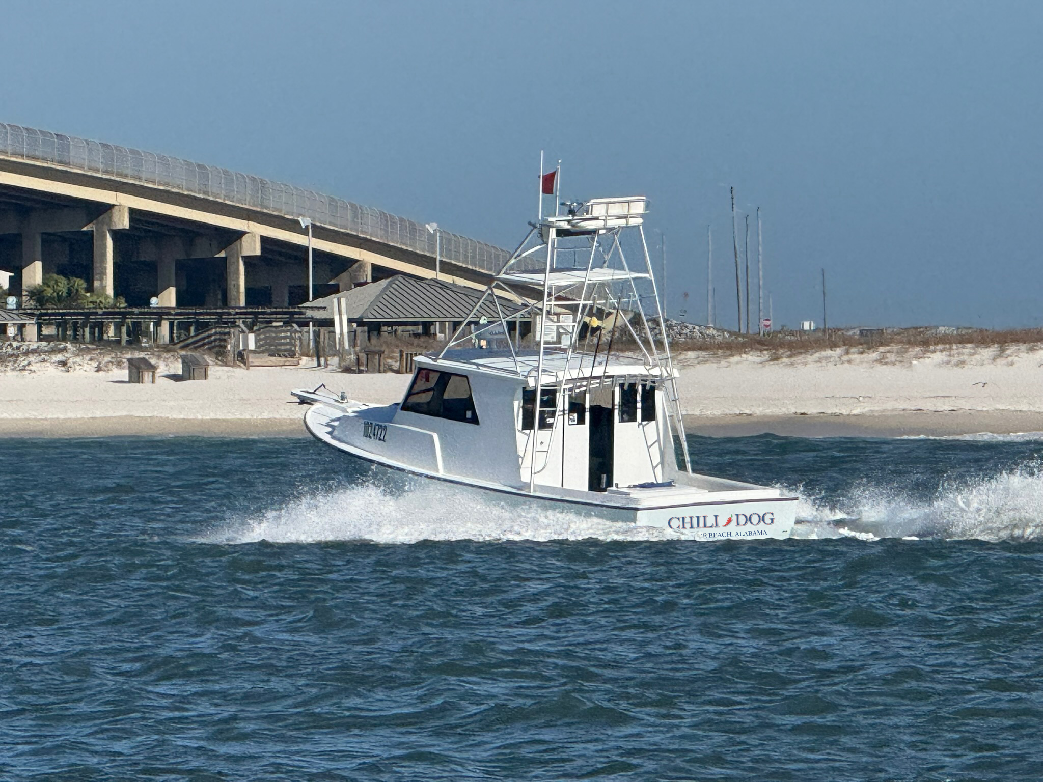 A white motorboat with the name 'Chili Dog' on the back, cruising through the water near a beach with sand dunes, a pavilion, and a bridge in the background, under a cloudy sky.