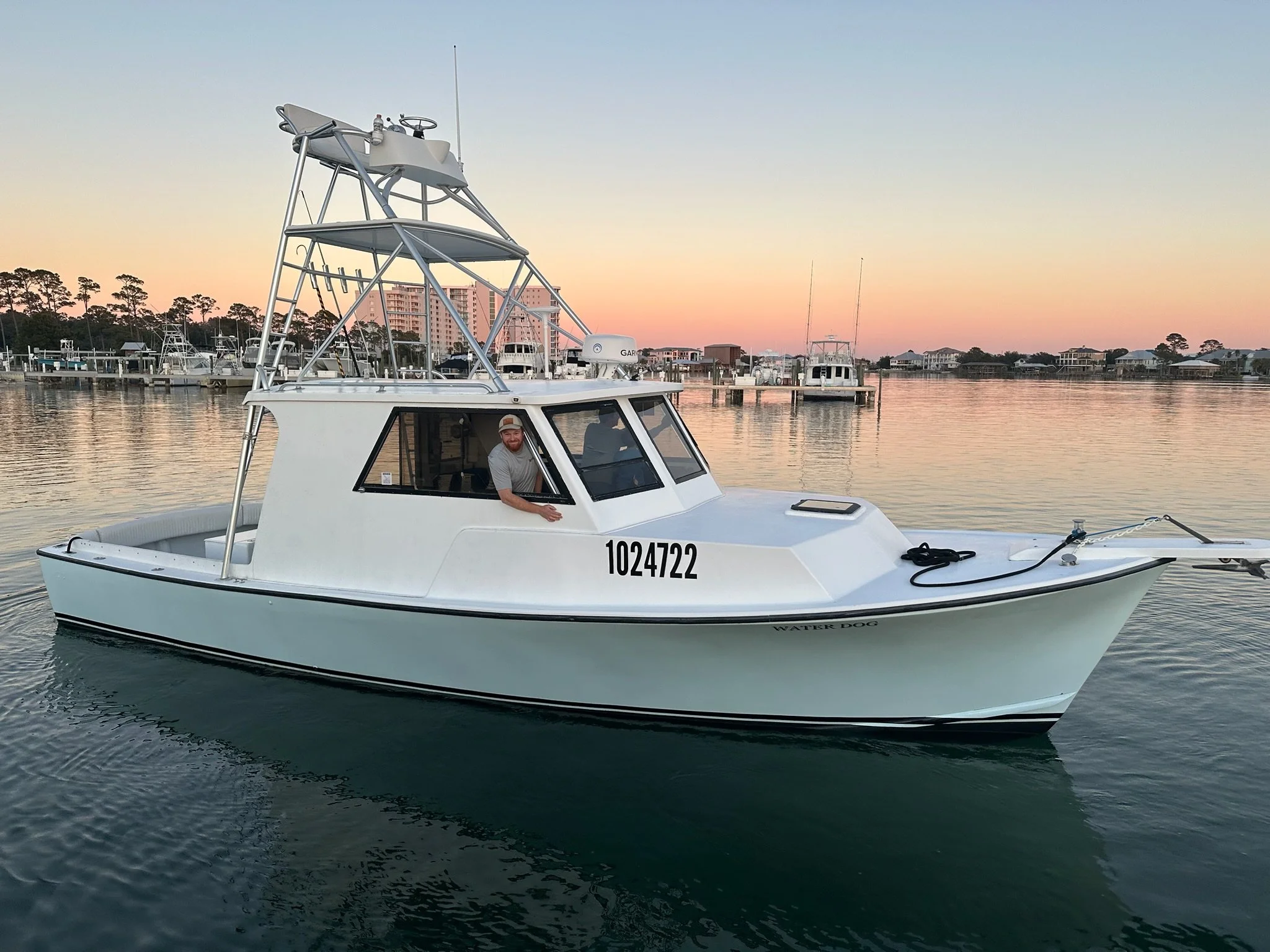 A man is sitting in the captain's seat of a white fishing boat named Water Dog, floating on calm water during sunset, with a marina and buildings in the background.